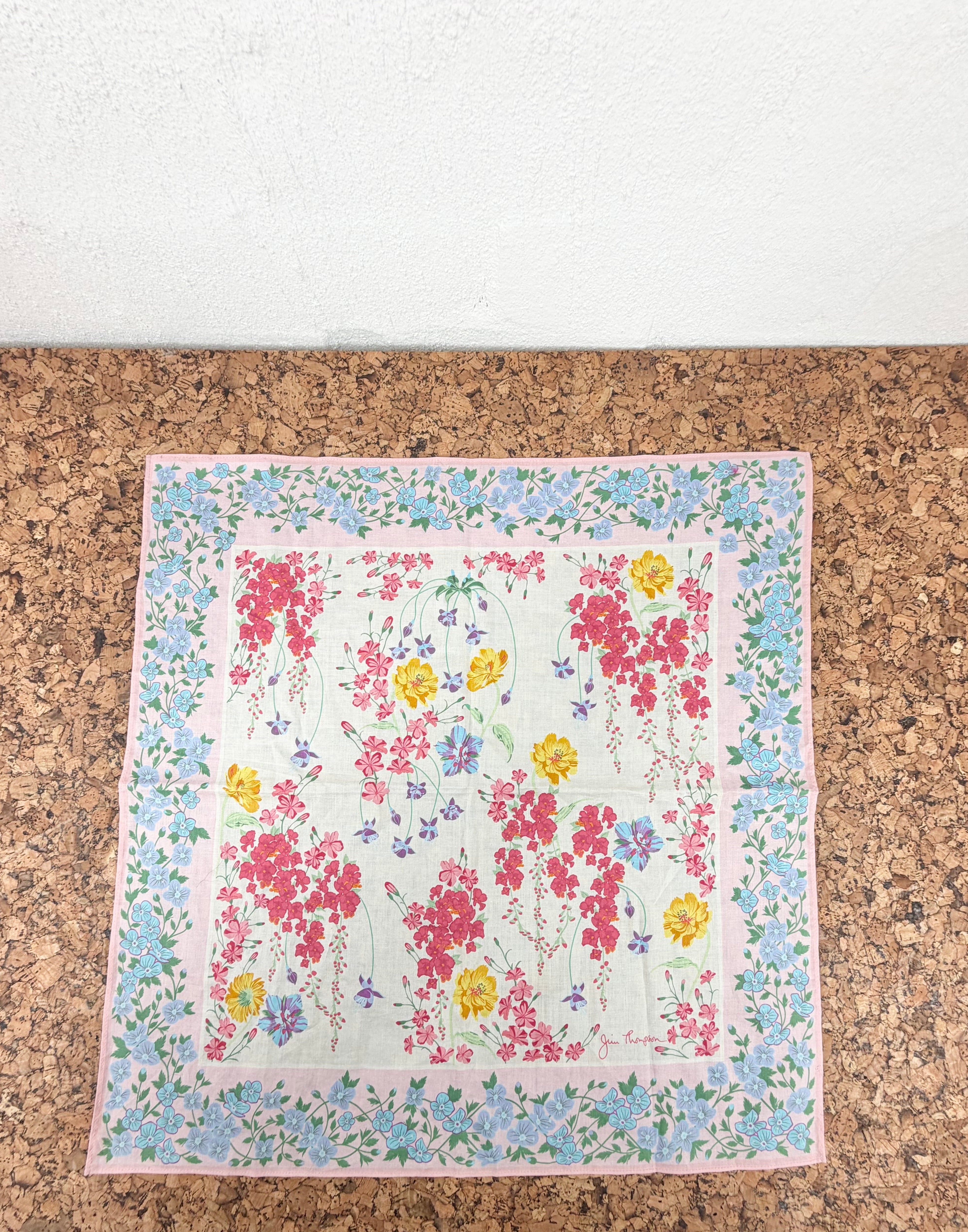 A square floral scarf with a pink border and a central white field featuring red, pink, yellow, and blue flowers, laid flat on a brown cork surface.