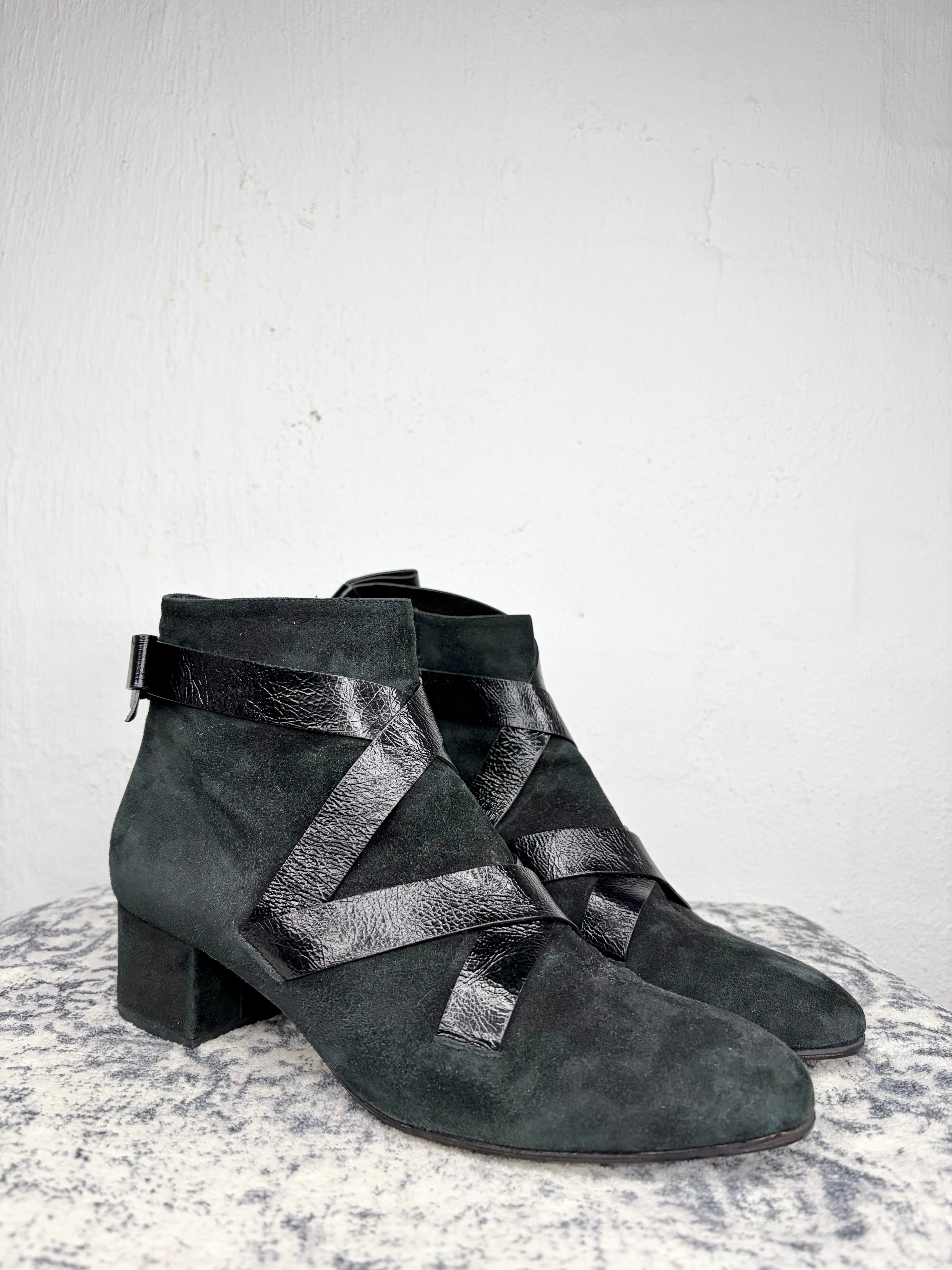 A pair of dark gray suede ankle boots with glossy black leather criss-cross straps and a low block heel, displayed on a textured gray surface against a white wall.