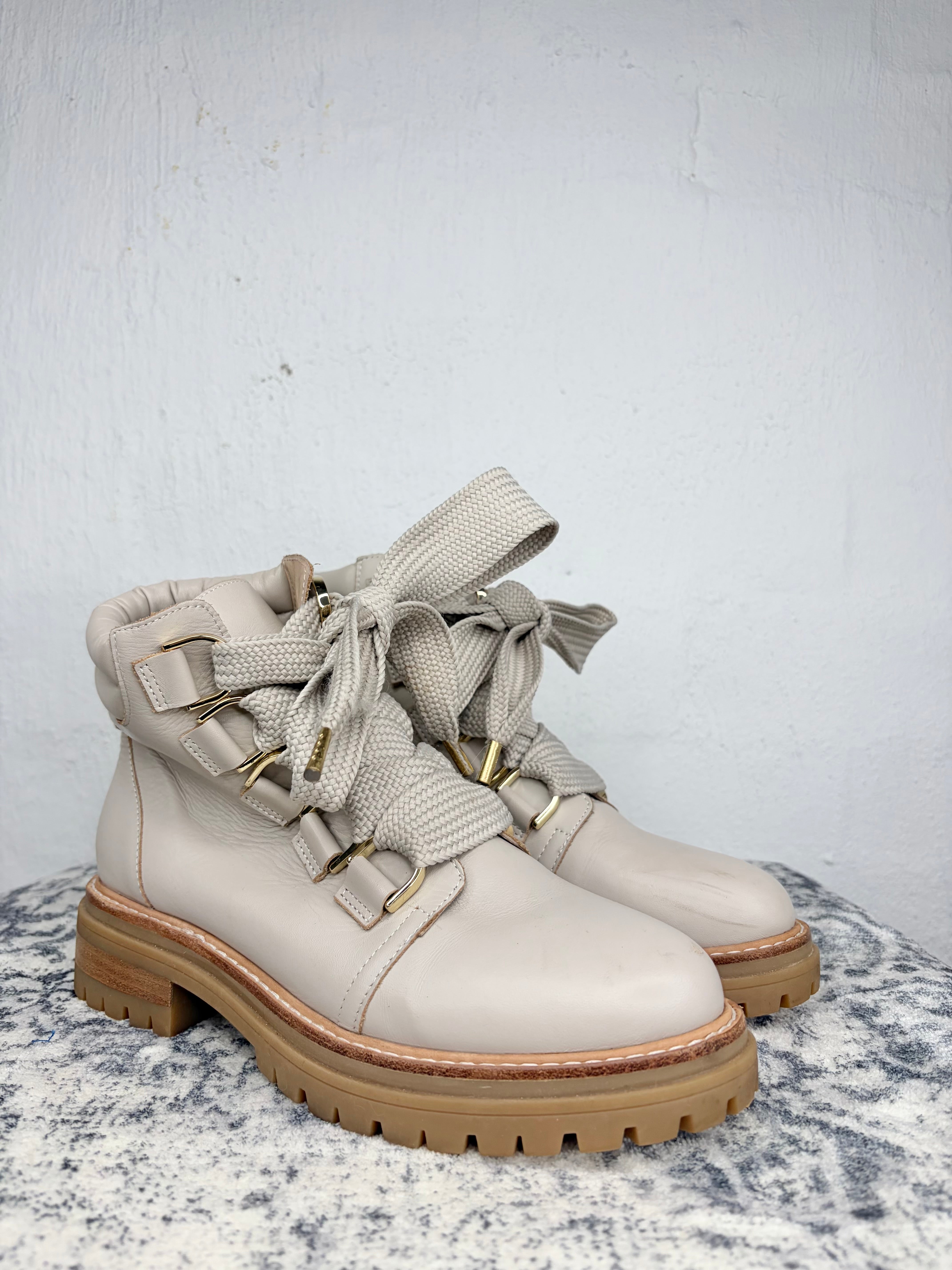 A pair of light beige leather lace-up ankle boots with wide fabric straps, gold-tone buckles, and chunky tan soles, displayed on a textured gray surface against a light gray wall.