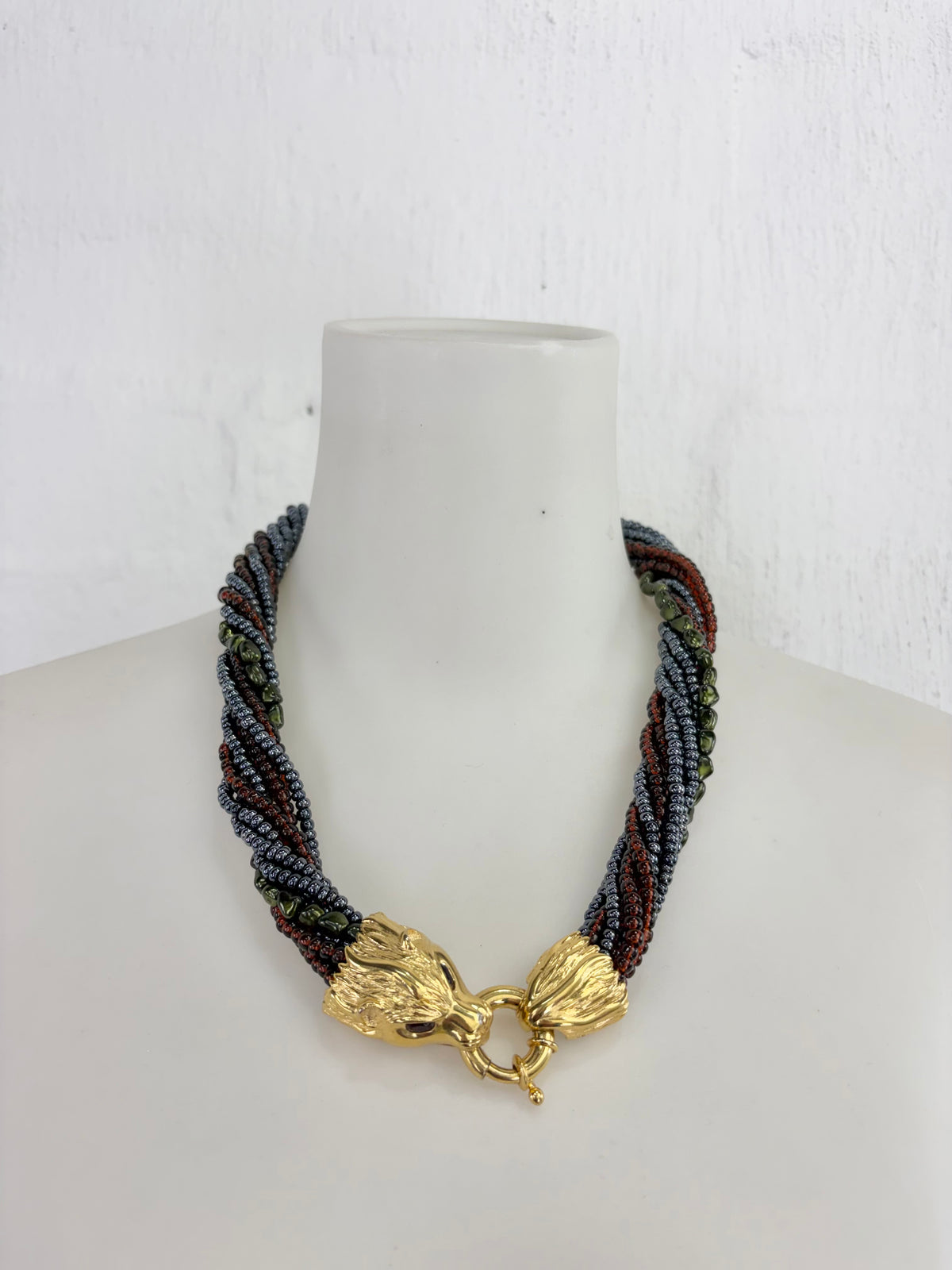 A multi-strand braided necklace with dark gray, brown, and green beads, featuring a textured gold clasp, displayed on a white mannequin bust.