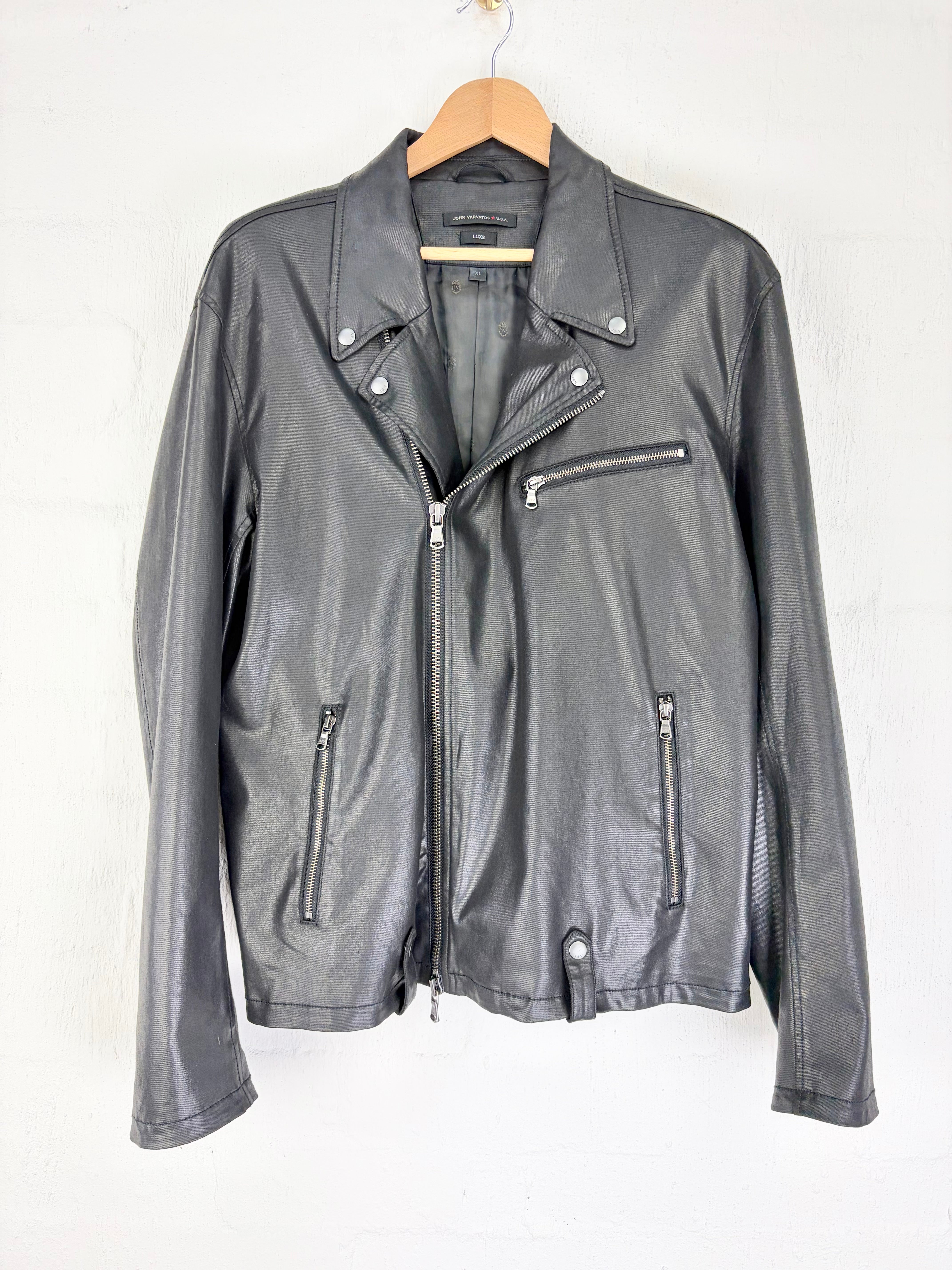 A black faux-leather biker jacket with silver zippers and snap buttons, hanging on a wooden hanger against a white wall.