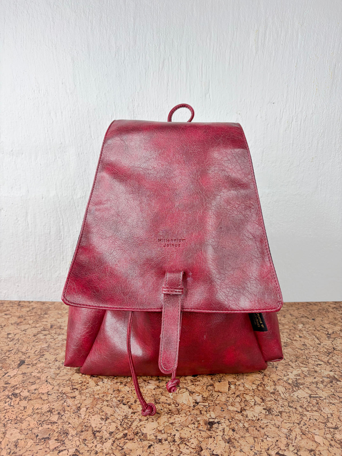 A distressed deep red leather backpack with a front flap, drawstring closure, and top loop handle, standing on a speckled brown countertop against a white wall.