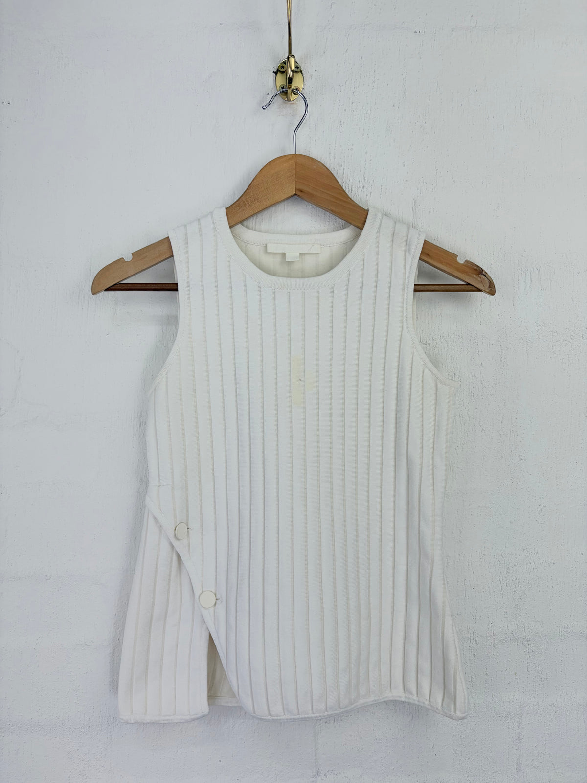 A sleeveless white ribbed knit vest with side buttons, hanging on a wooden hanger against a white textured wall.