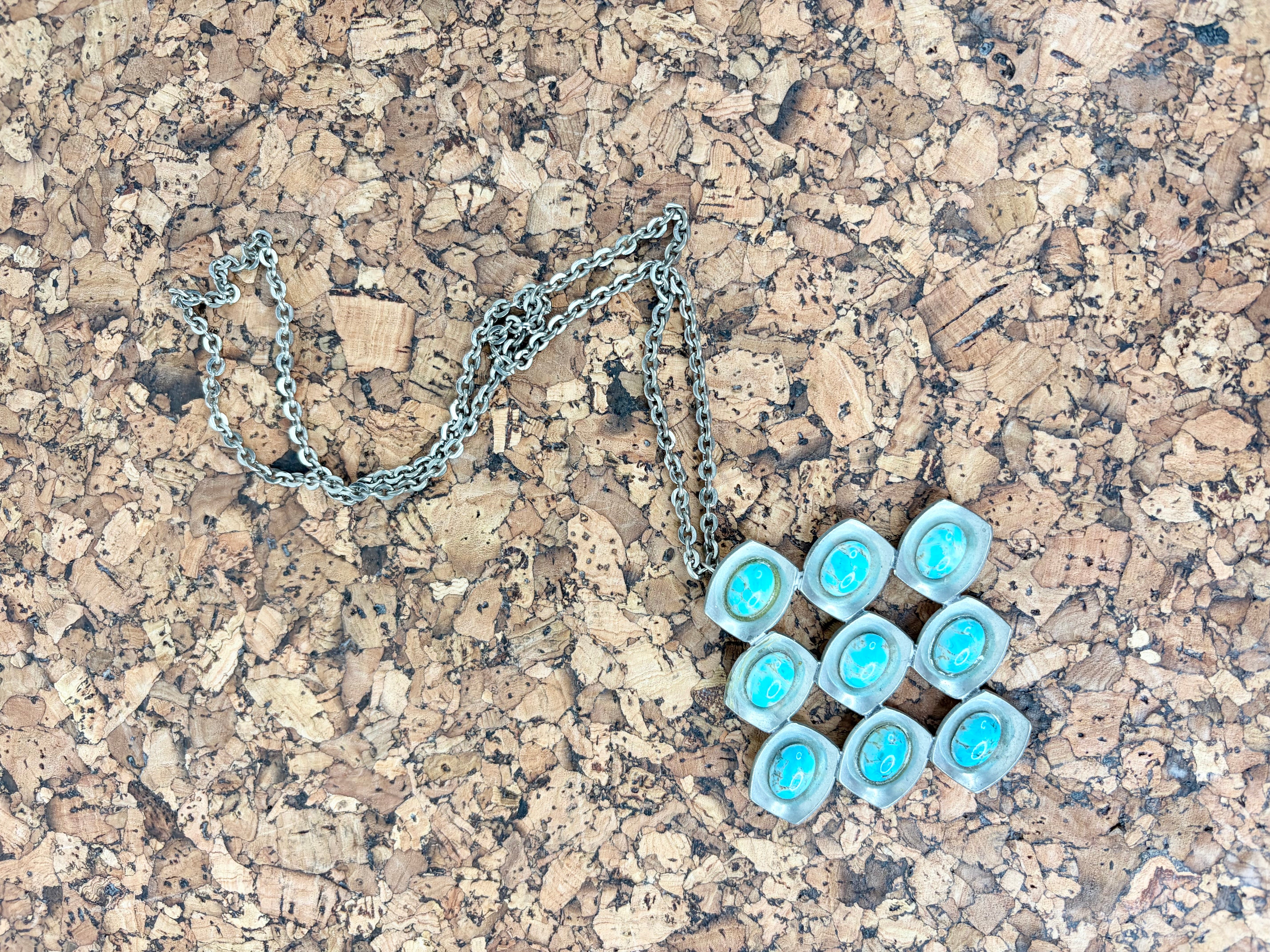 A silver chain necklace with a pendant made of nine small turquoise-blue oval stones set in silver, displayed on a cork surface.