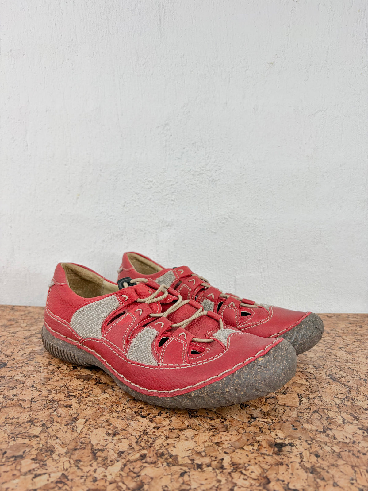 A pair of red outdoor slip-on shoes with beige laces and textured gray soles, resting on a cork mat against a white wall.
