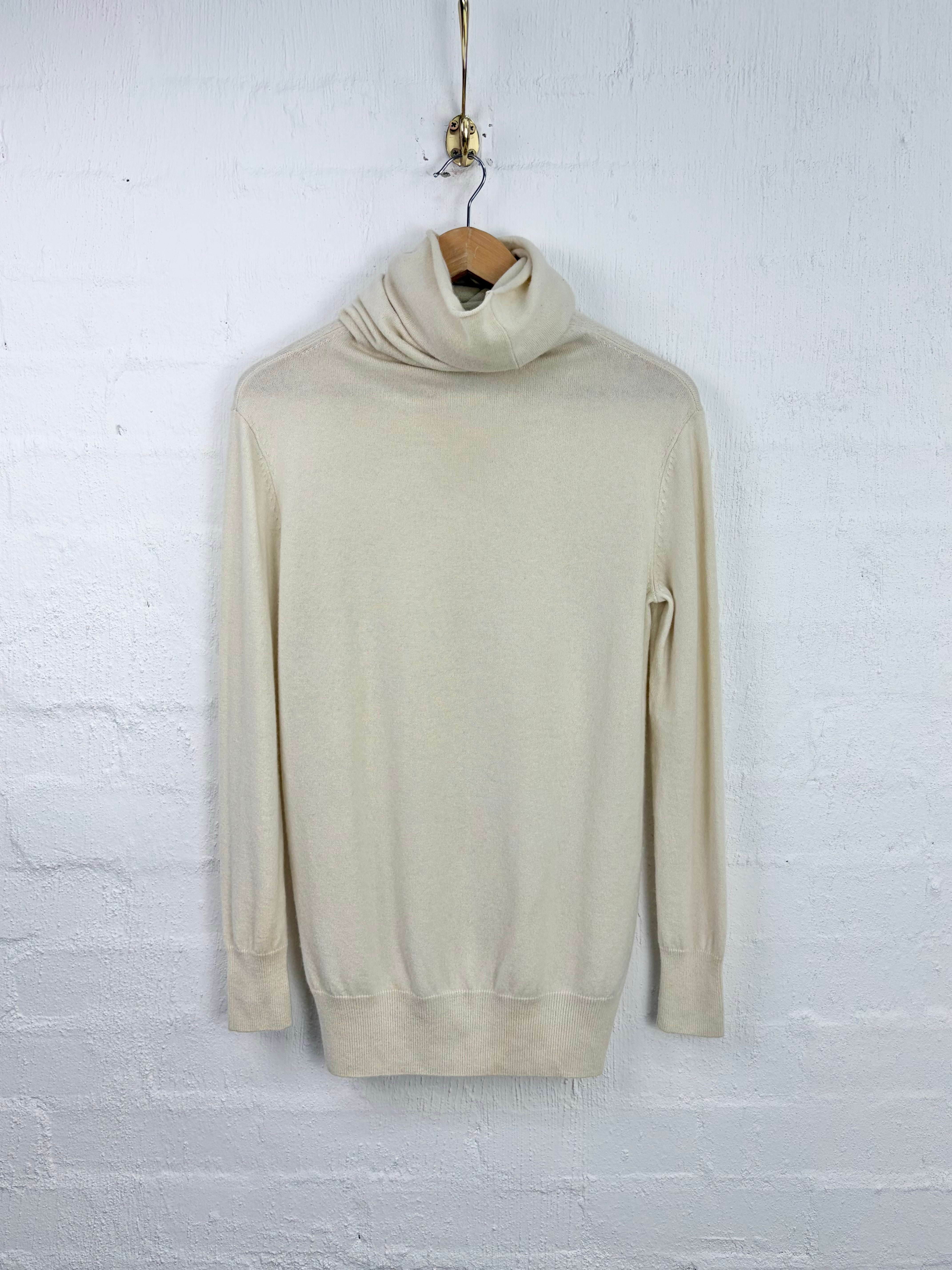 A cream-colored knit turtleneck sweater hanging on a wooden hanger against a white painted brick wall.