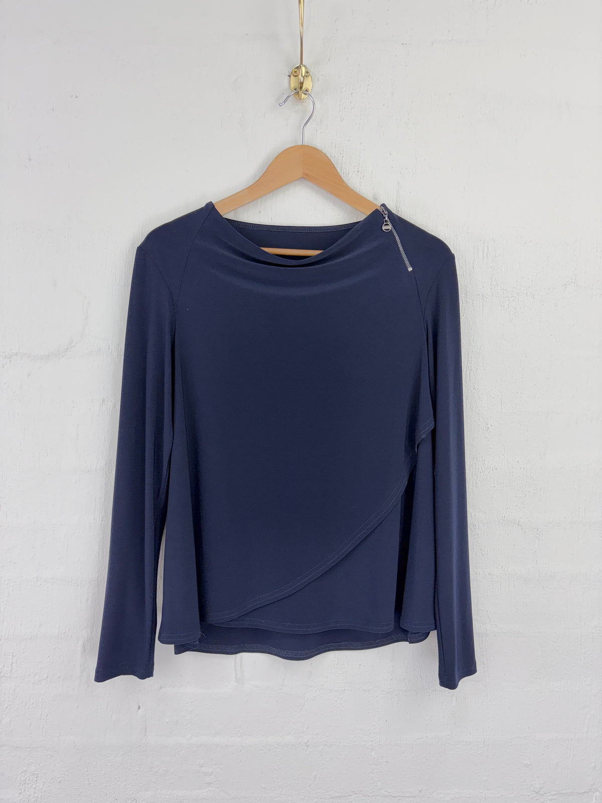 A navy blue long-sleeve top with an asymmetrical wrap-style hem and a small silver zipper detail on the shoulder, hanging on a wooden hanger against a white brick wall.