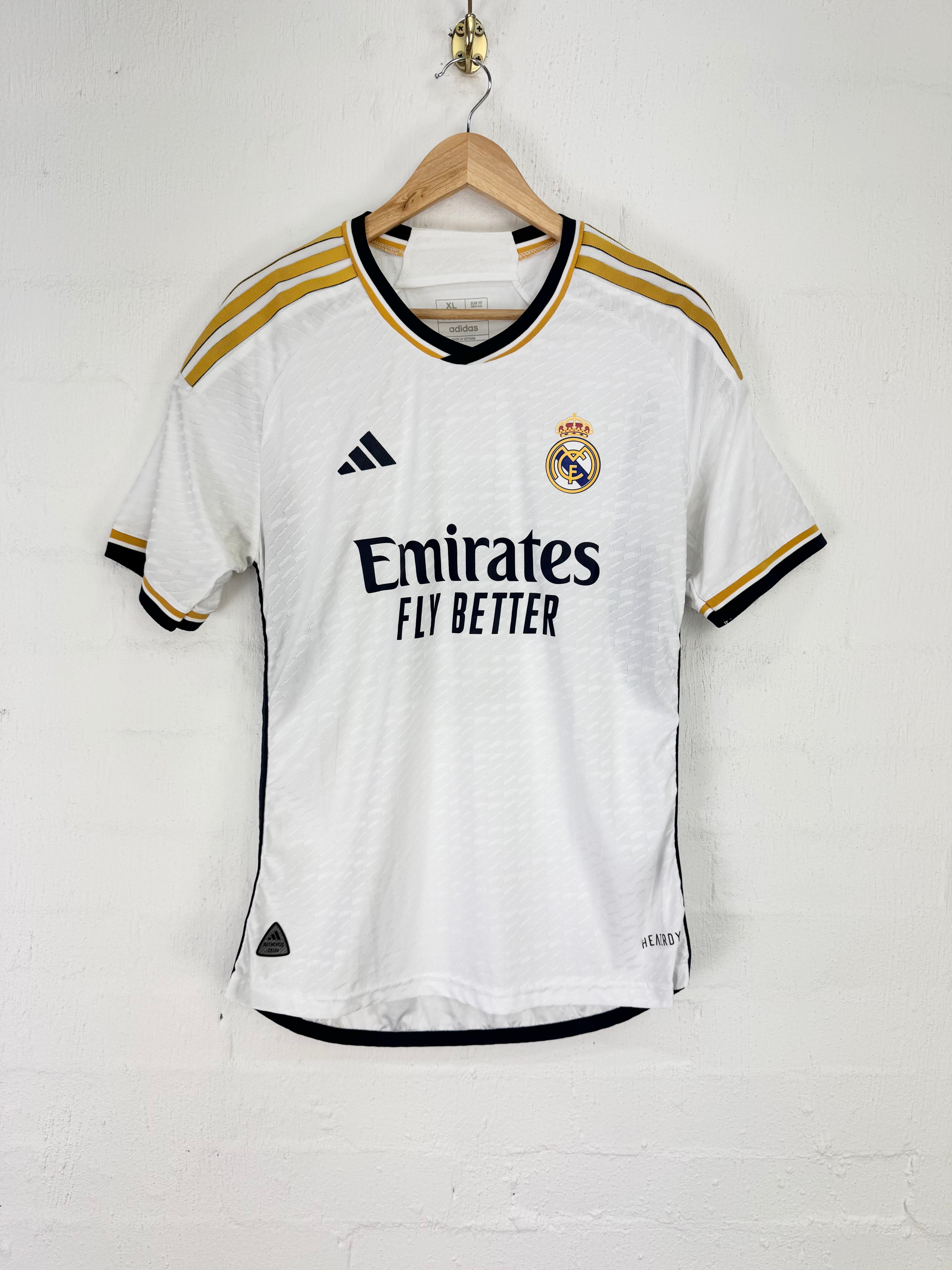 A white Real Madrid soccer jersey by Adidas, featuring gold and black stripes on the shoulders and collar, with the Emirates Fly Better sponsor logo on the chest, hanging on a wooden hanger against a white brick wall.