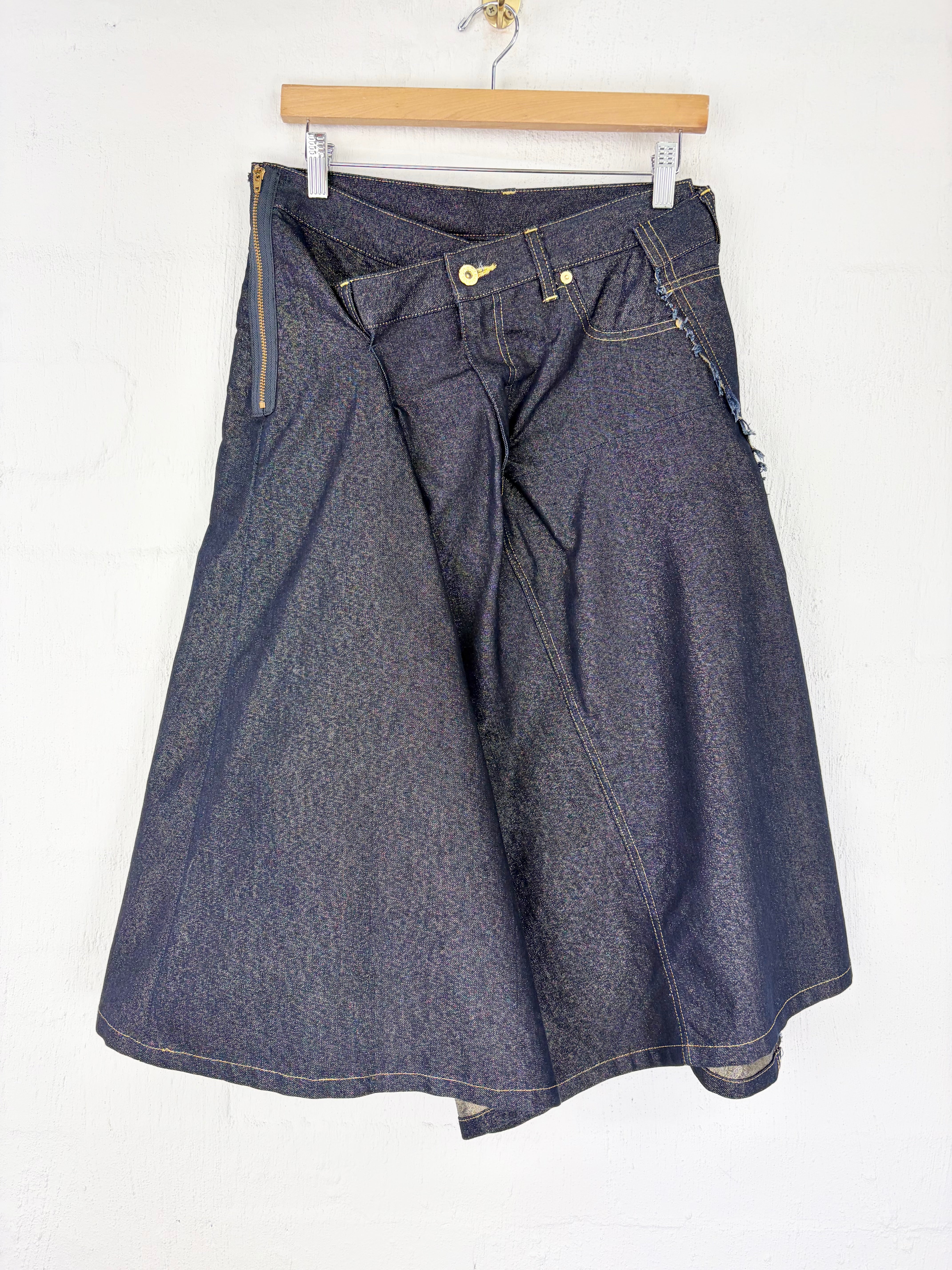 A dark blue denim skirt with an asymmetrical wrap design, hanging on a wooden hanger against a white wall.
