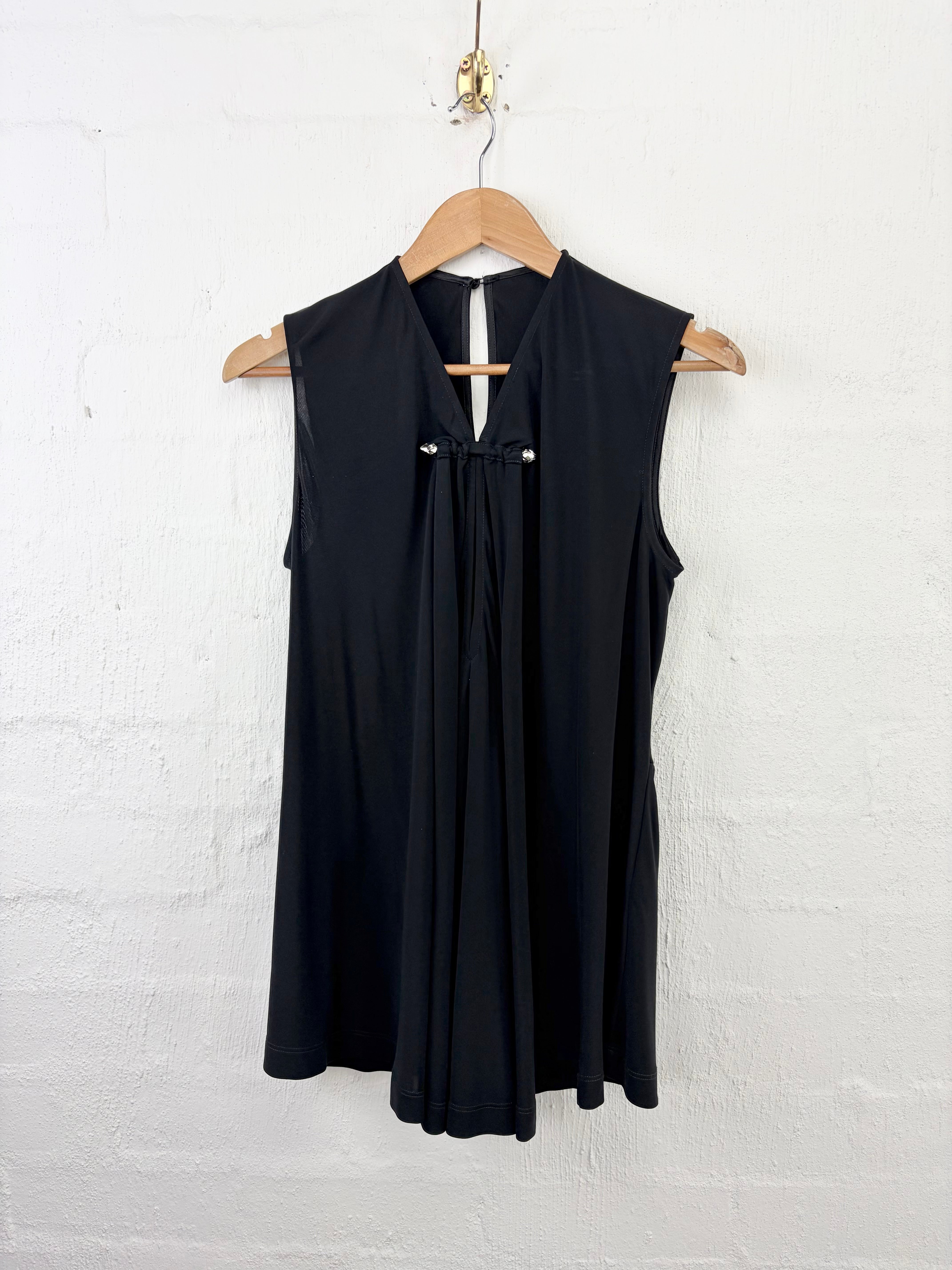 A sleeveless black dress with a keyhole back and a gathered front tie detail, hanging on a wooden hanger against a white painted brick wall.