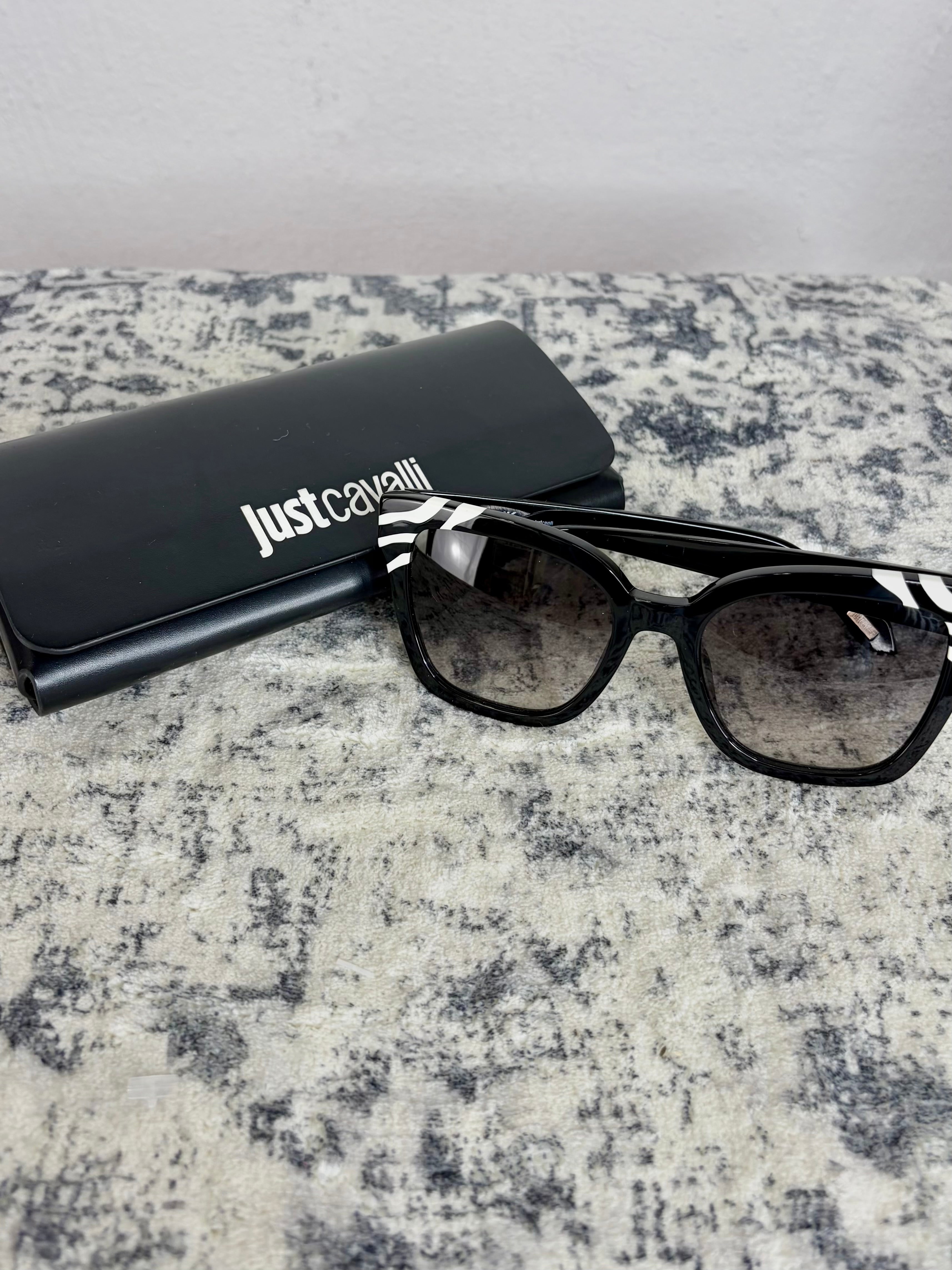 Black cat-eye sunglasses with a black and white zebra-patterned temple arm, shown next to a black Just Cavalli case on a textured gray surface.