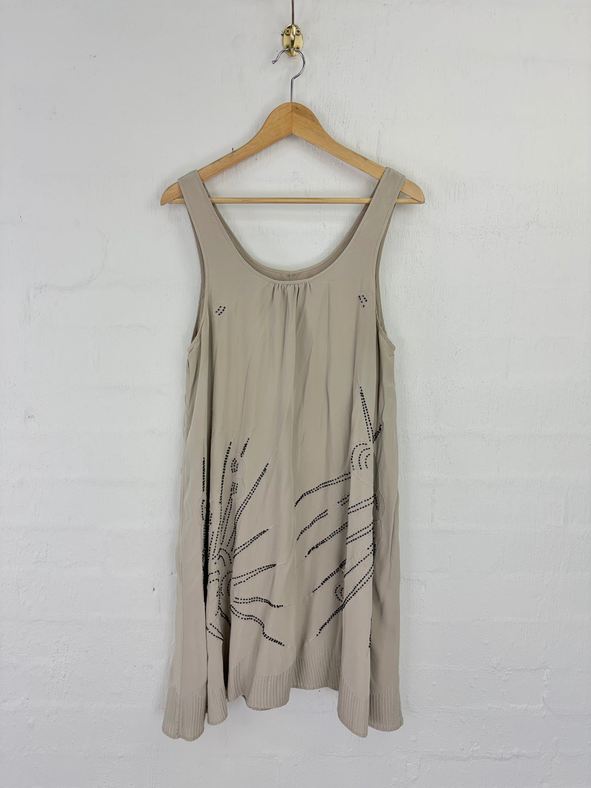 A sleeveless beige tank top with black line-art sunburst graphics on the lower front, hanging on a wooden hanger against a white painted brick wall.