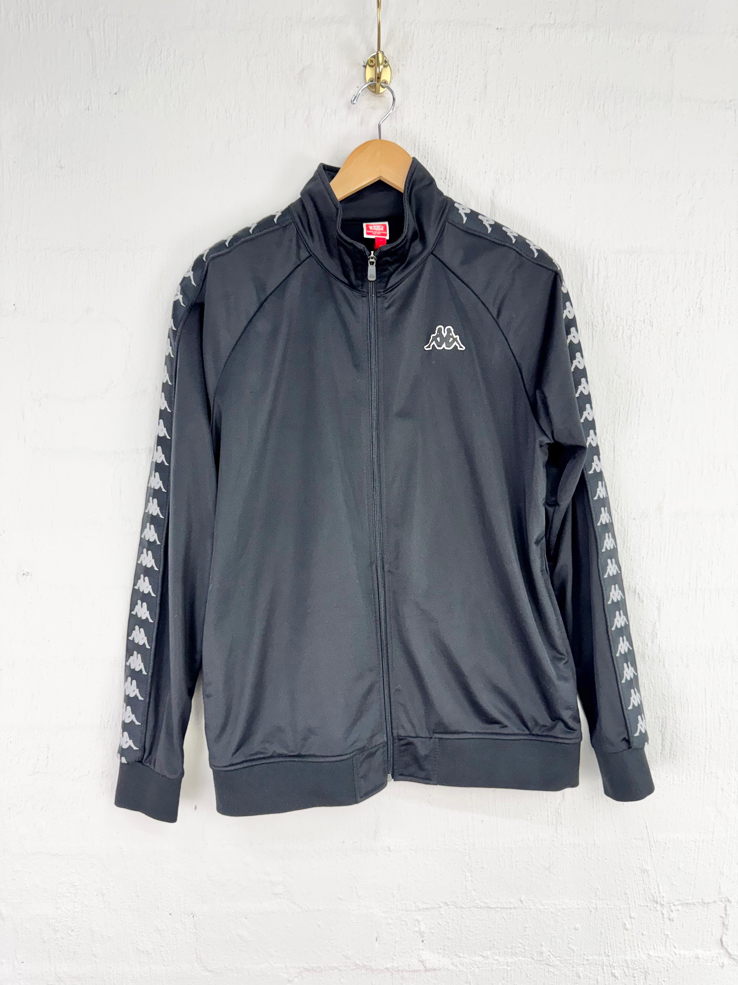 A dark gray Kappa track jacket hanging on a wooden hanger against a white brick wall. The jacket features the signature Kappa logo on the left chest and a repeating Kappa horse pattern down both sleeves.
