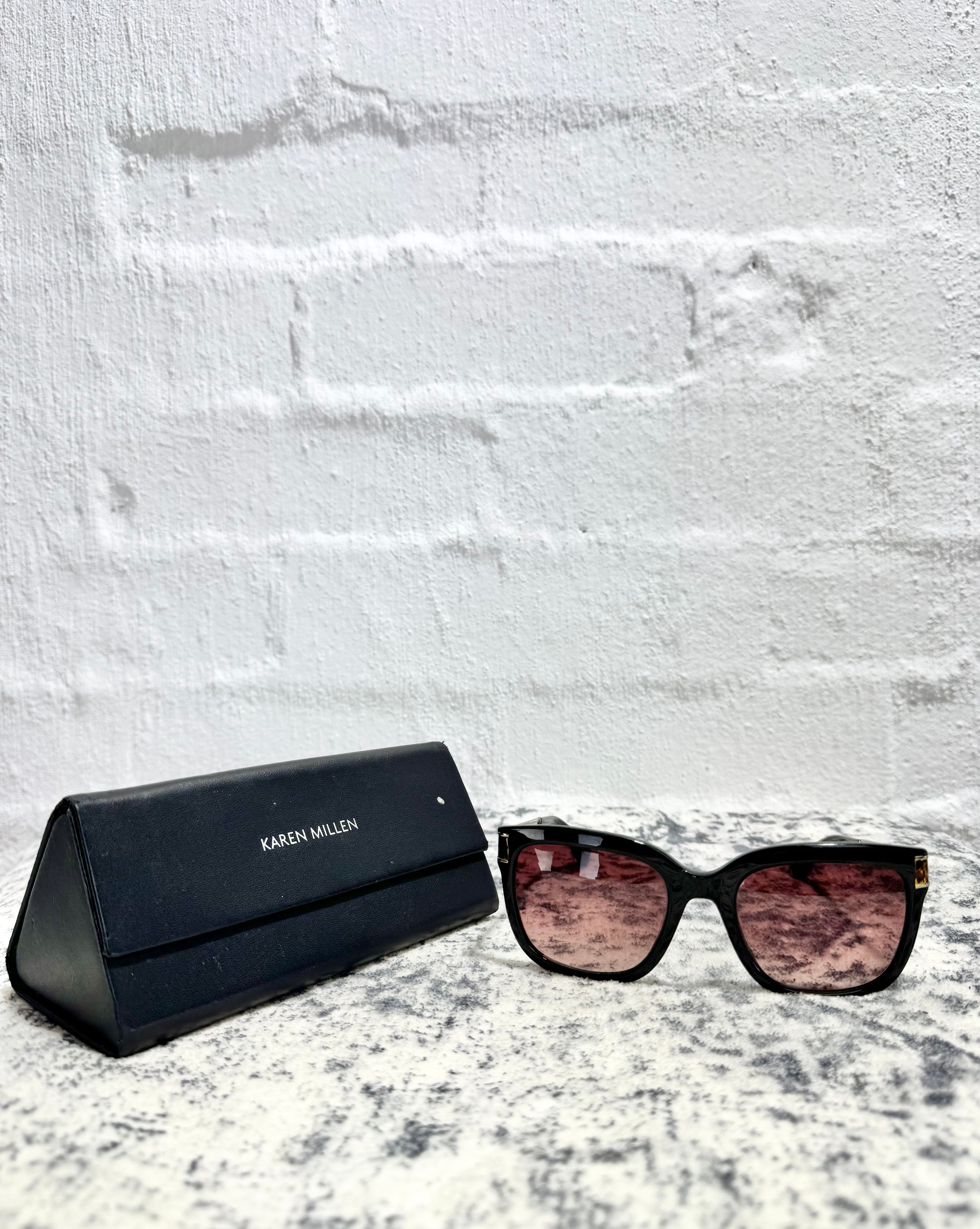 A pair of black cat-eye sunglasses with pink-tinted lenses next to a black KAREN MILLEN brand case, set on a speckled white and gray surface against a white brick wall.