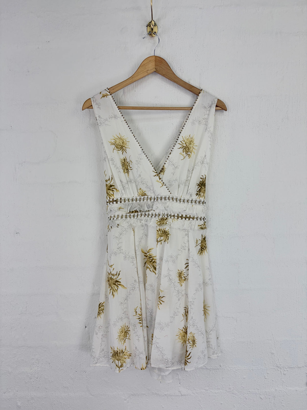 A sleeveless white wrap-style dress with a gold floral print and embroidered detailing, hanging on a wooden hanger against a white brick wall.