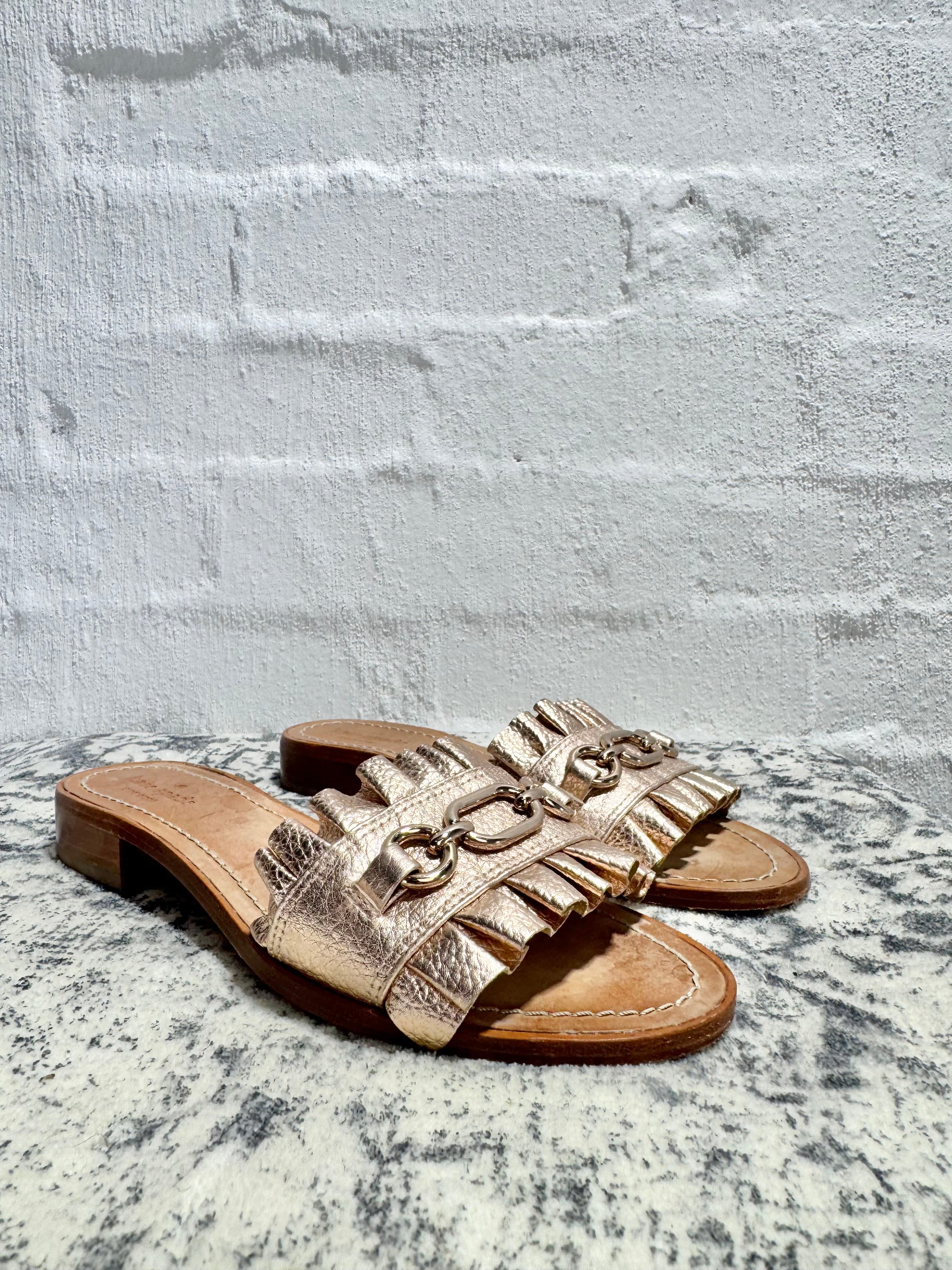 A pair of metallic rose gold slide sandals with fringed straps and gold-tone chain details, resting on a speckled stone surface against a white painted brick wall.