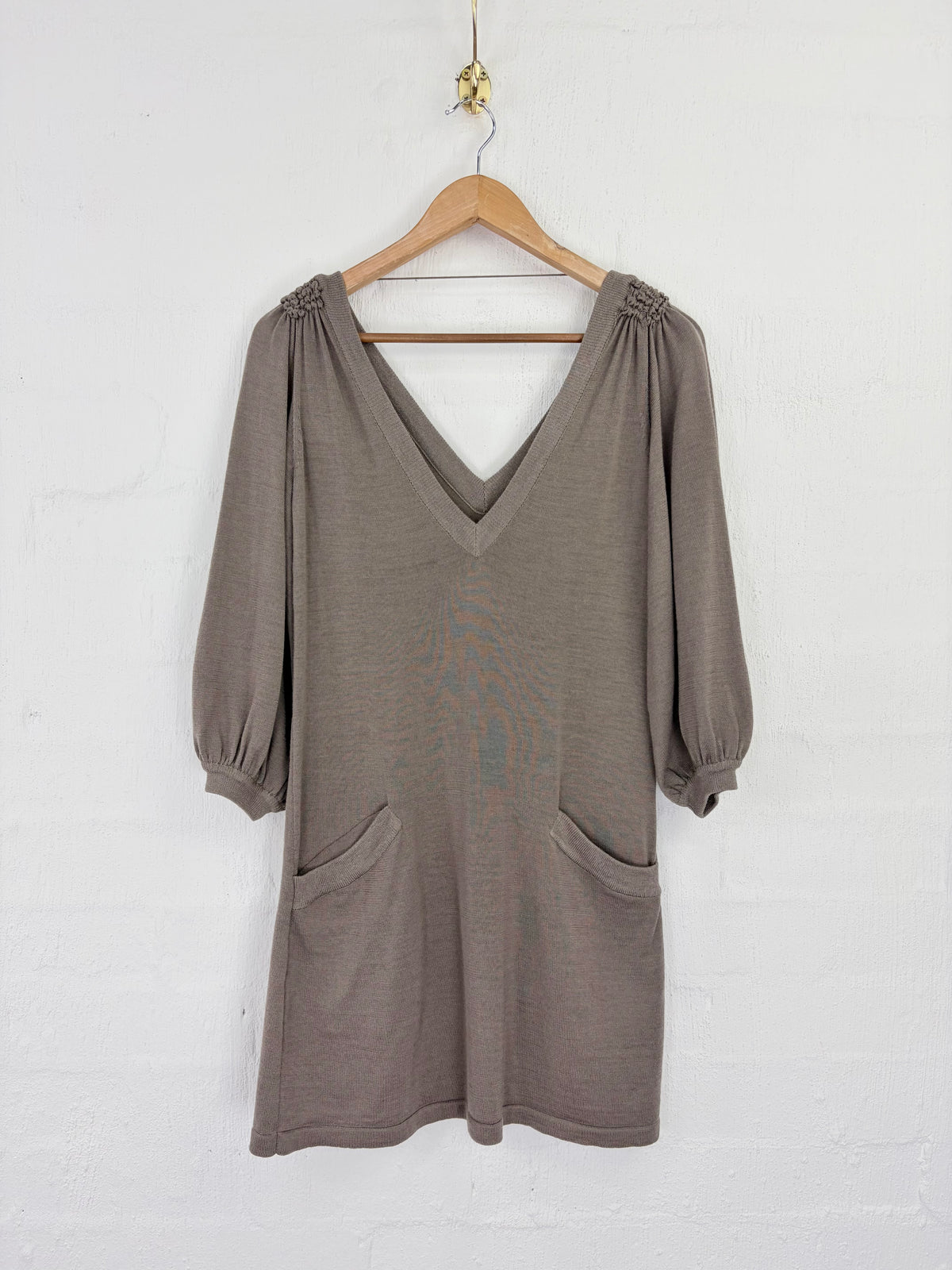 A taupe brown knit dress with three-quarter balloon sleeves and gathered shoulder details, hanging on a wooden hanger against a white painted brick wall.