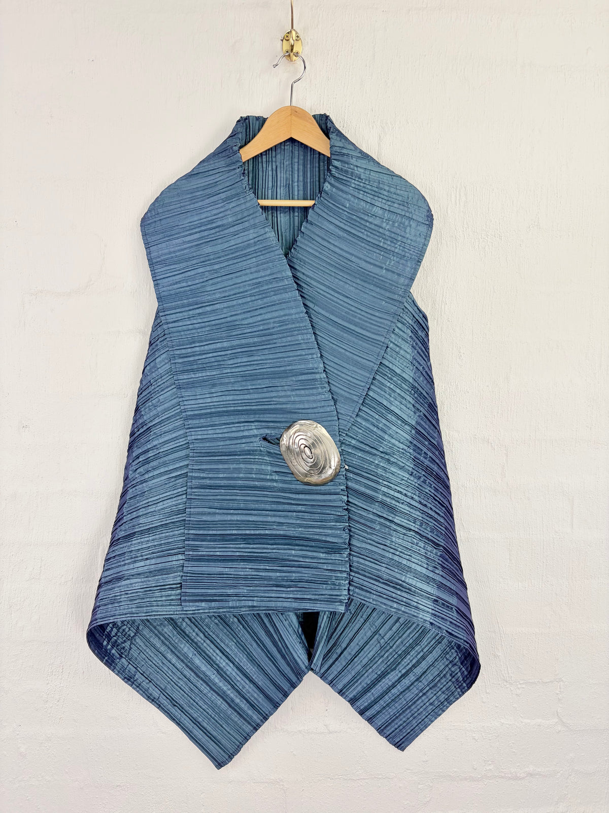 A blue striped wrap vest with a large silver spiral button, hanging on a wooden hanger against a white painted brick wall.