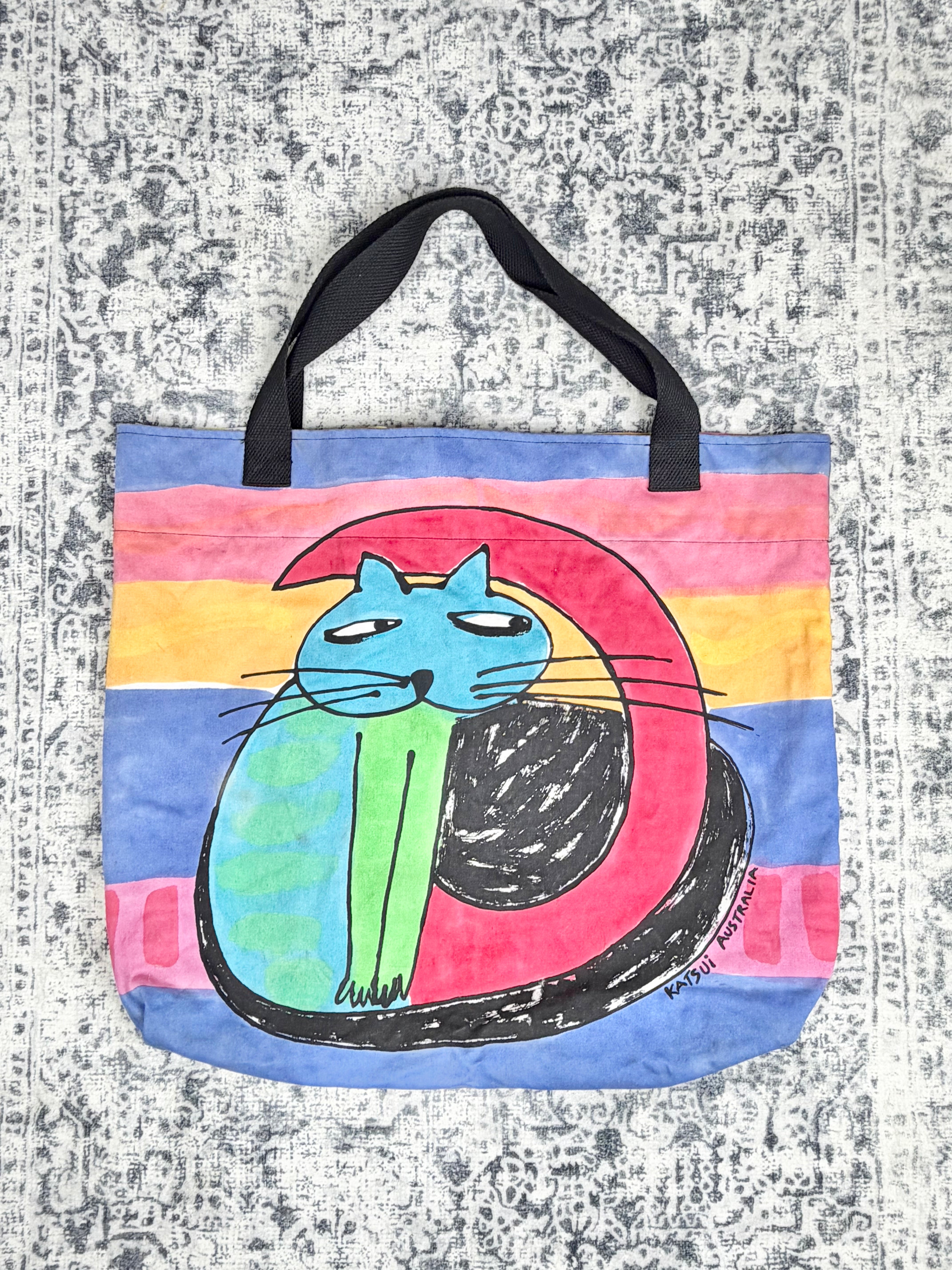A canvas tote bag featuring a colorful hand-drawn illustration of a blue cat inside a pink and green yin-yang symbol, set against a background of pink, orange, yellow, and blue stripes. The bag has black handles and is photographed flat on a textured gray and white patterned surface.