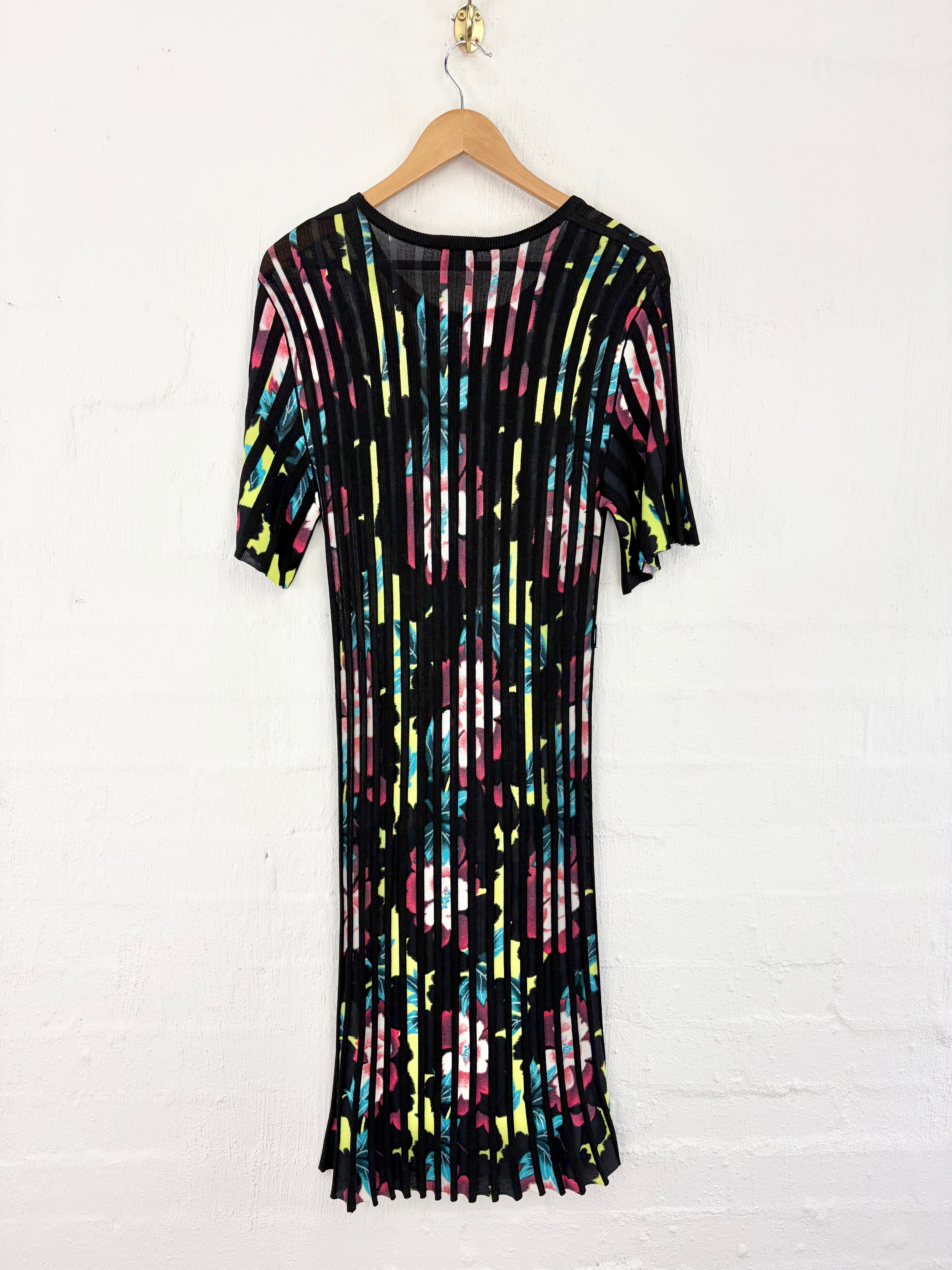 A short-sleeve black mesh dress with a vibrant multicolor vertical stripe pattern, hanging on a wooden hanger against a white painted brick wall.
