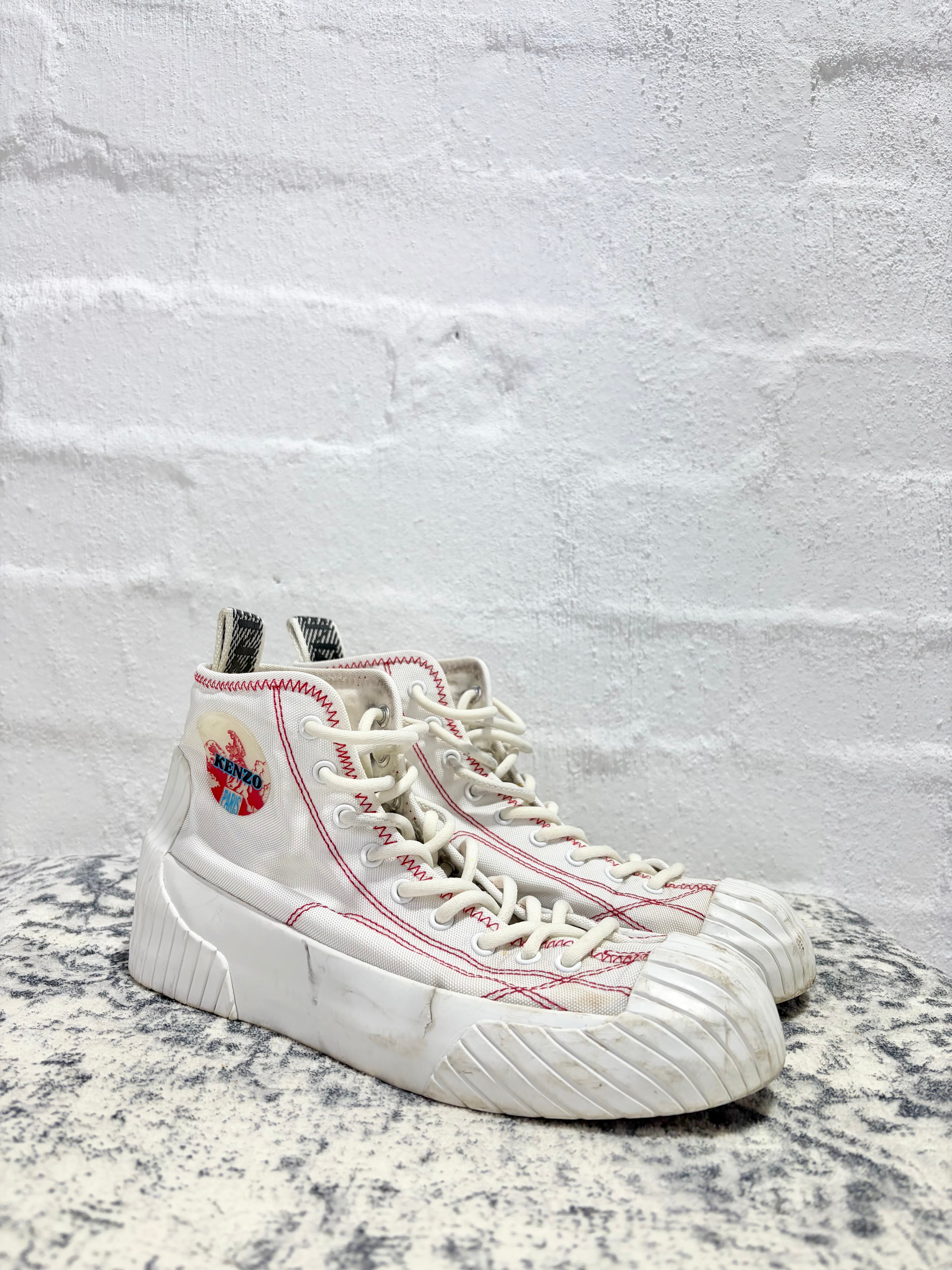 A pair of high-top white sneakers with red contrast stitching, a red and blue graphic patch on the side, and chunky white soles, displayed on a speckled surface against a white brick wall.