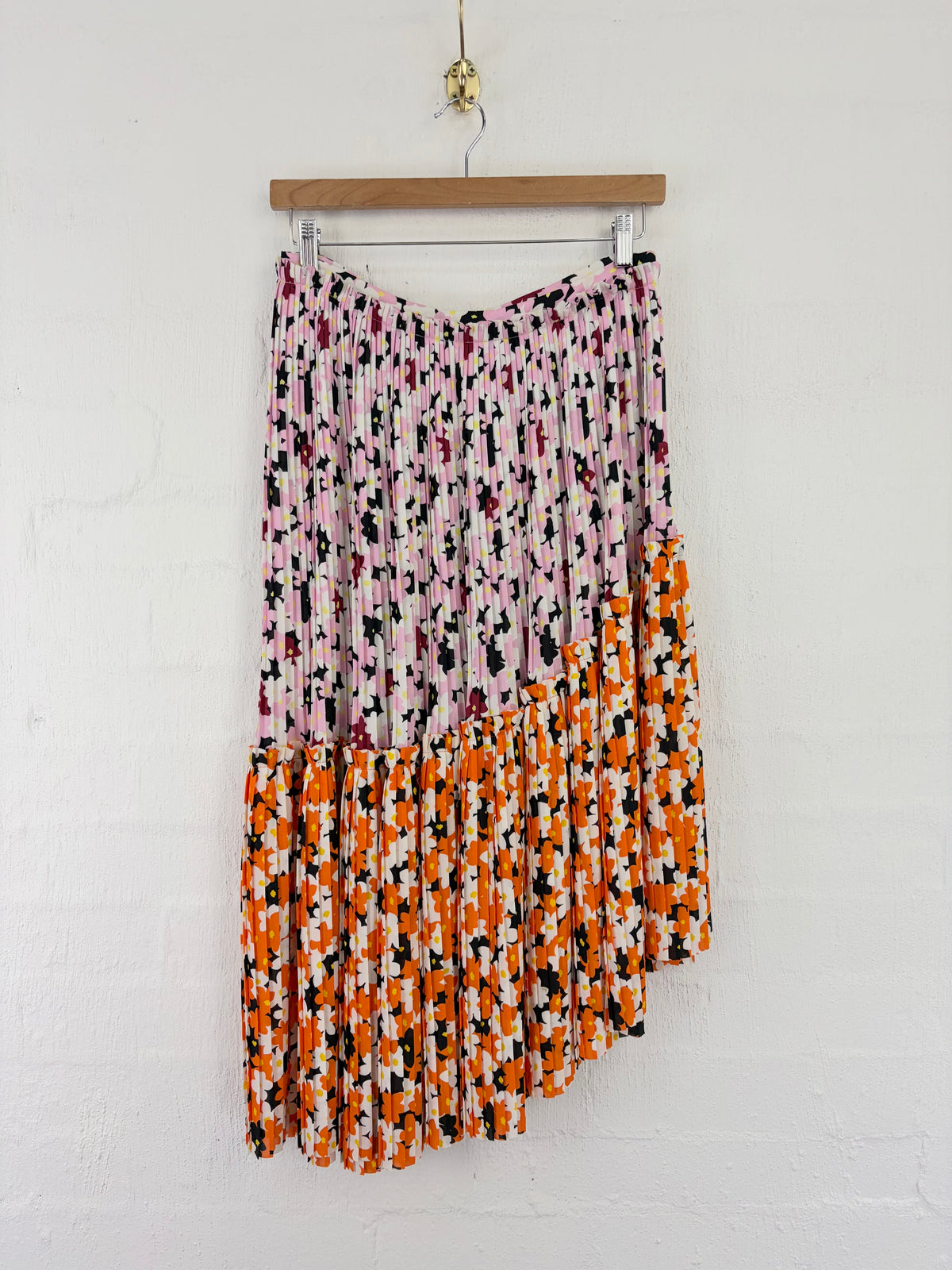 A pleated asymmetrical midi skirt hanging from a wooden hanger against a white painted brick wall. The top half of the skirt is light pink with a black, white, and magenta abstract print, while the bottom half is orange with a matching abstract print.