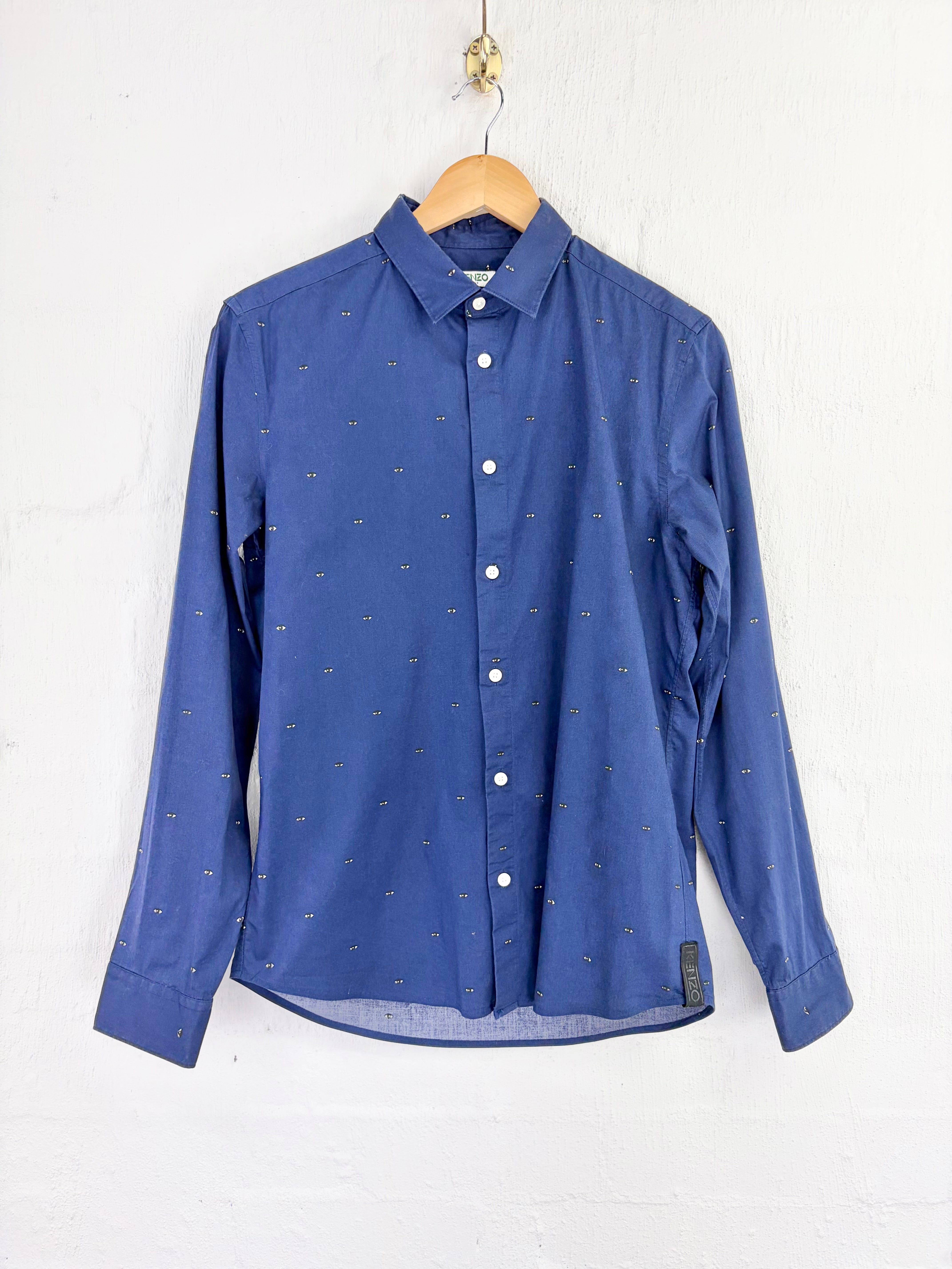 A blue long-sleeve button-up shirt with a tiny repeating cross pattern, hanging on a wooden hanger against a white painted brick wall.