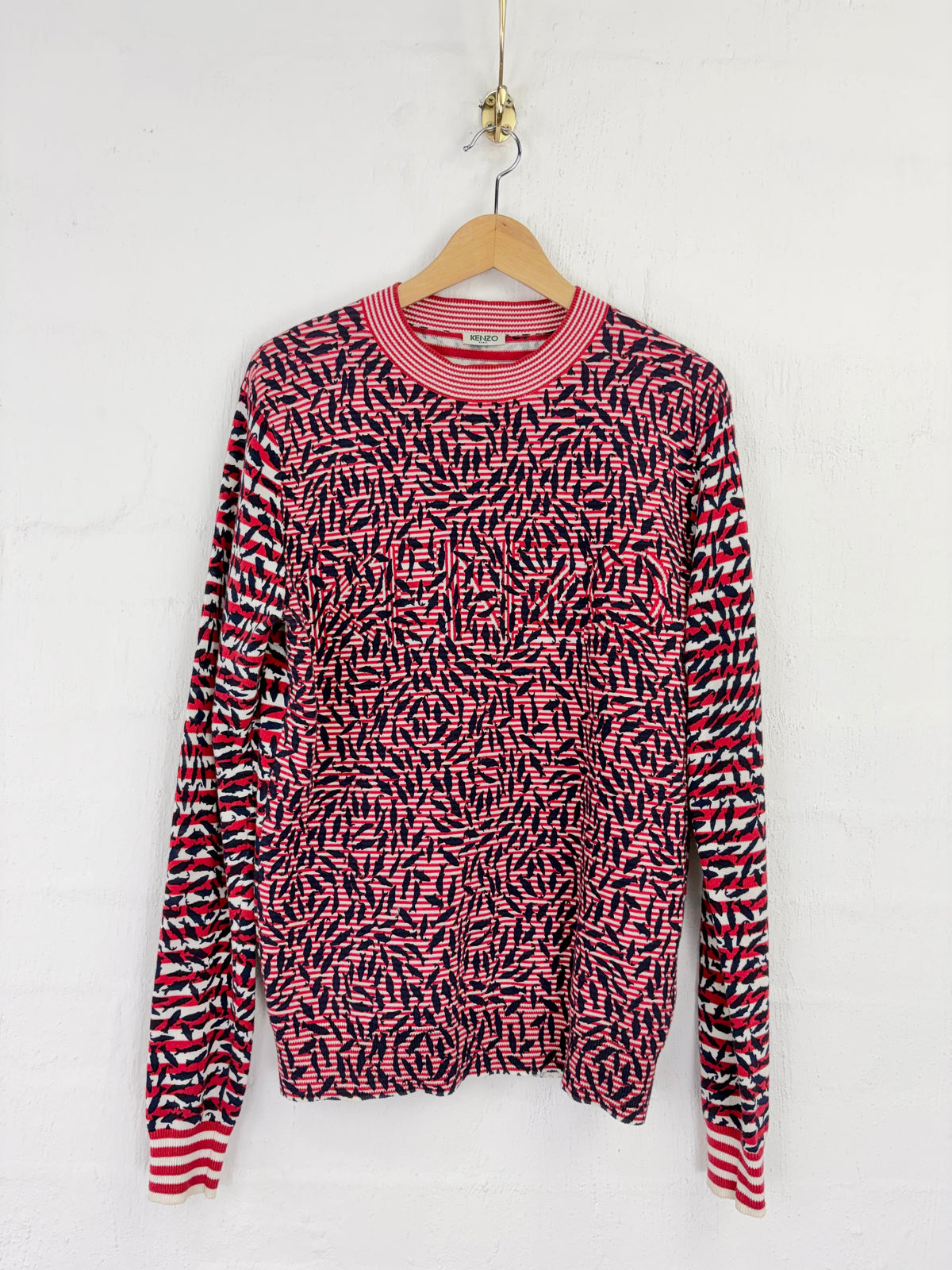 A pink and black patterned knit sweater with red and white striped cuffs and collar, hanging on a wooden hanger against a white wall.