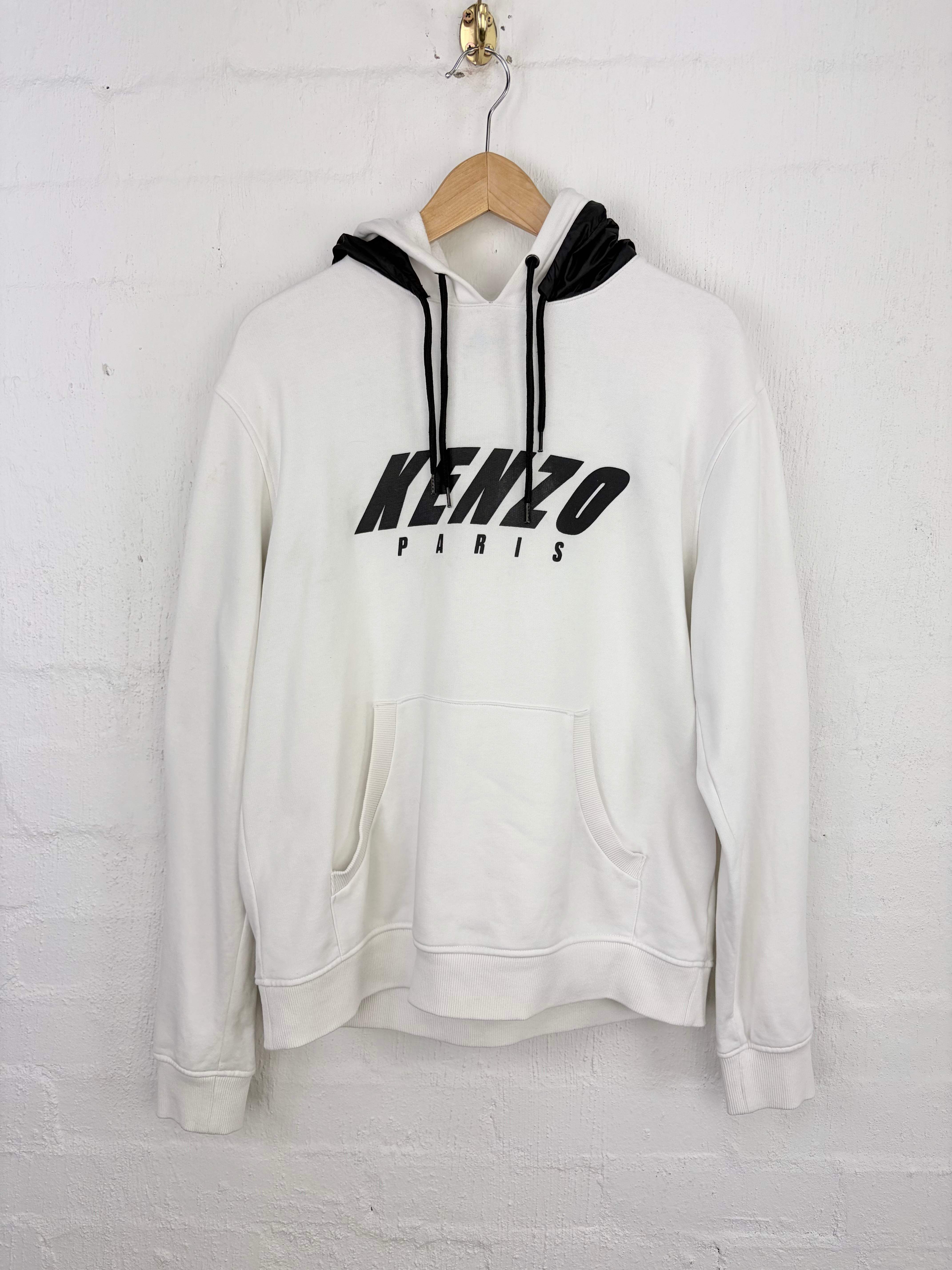 A white Kenzo Paris pullover hoodie with black drawstrings and a black logo on the chest, hanging on a wooden hanger against a white brick wall.