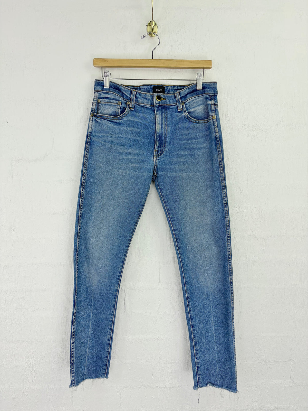 Light blue denim jeans with a raw, frayed hem, hanging from a wooden hanger against a white painted brick wall.