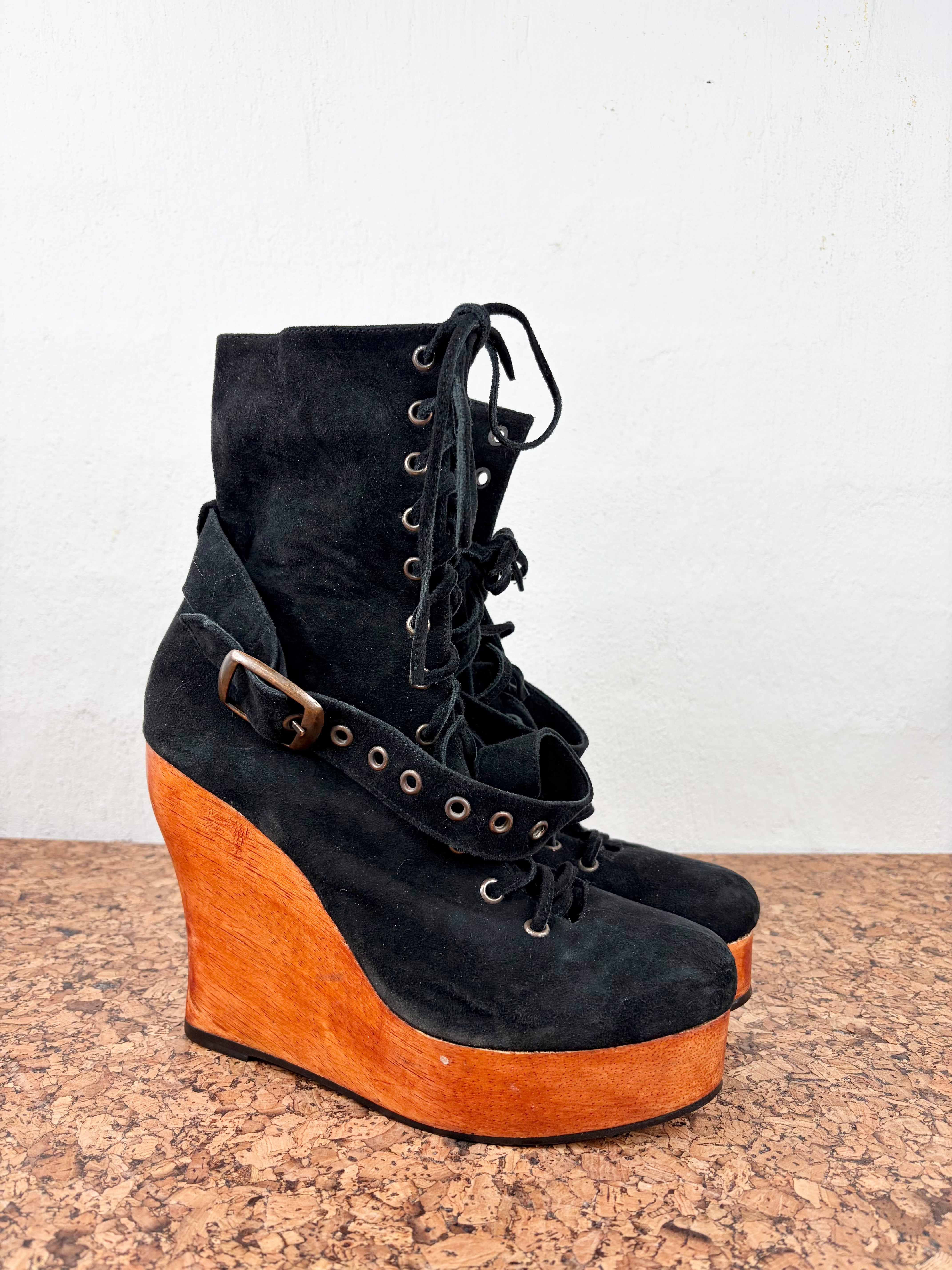 A pair of black suede lace-up wedge ankle boots with a thick orange-brown wooden platform wedge and a decorative buckle strap, displayed on a speckled brown surface against a white wall.