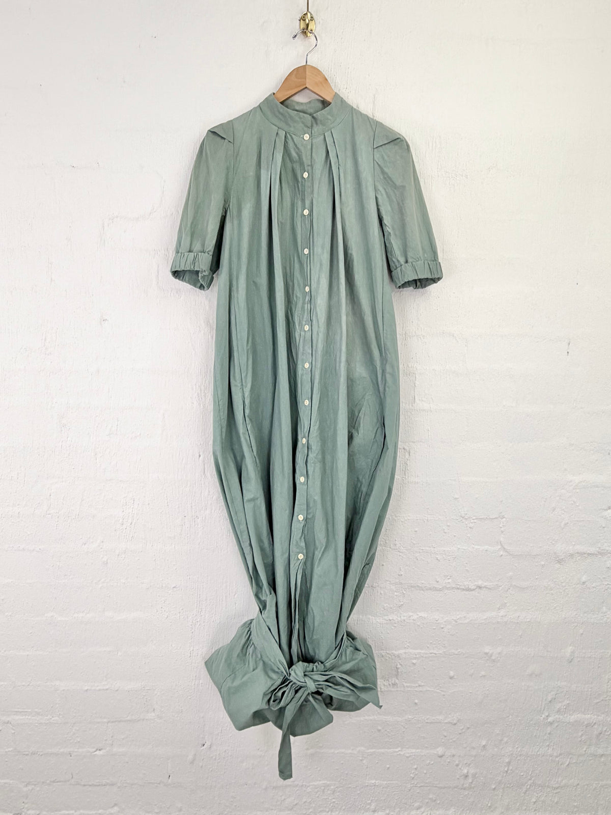 A sage green short-sleeve button-front dress with a gathered, balloon-like silhouette and a knotted hem, hanging on a wooden hanger against a white painted brick wall.