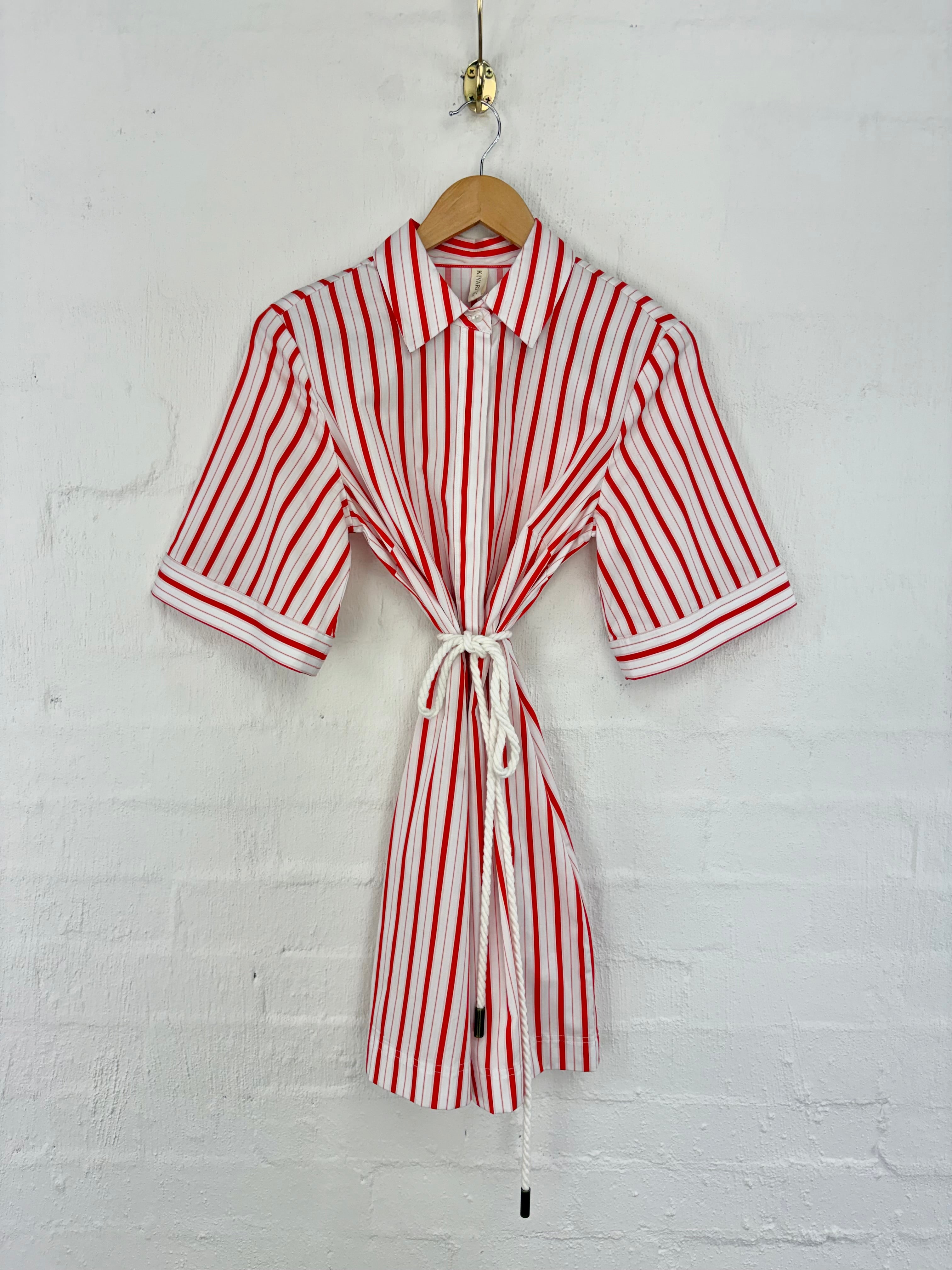 A red and white vertically striped short-sleeve shirt dress with a white rope tie belt, hanging on a wooden hanger against a white painted brick wall.