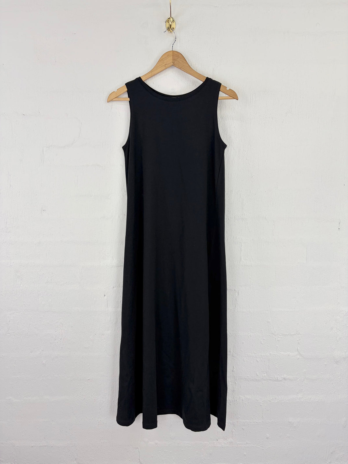 A sleeveless black midi dress hanging on a wooden hanger against a white painted brick wall.