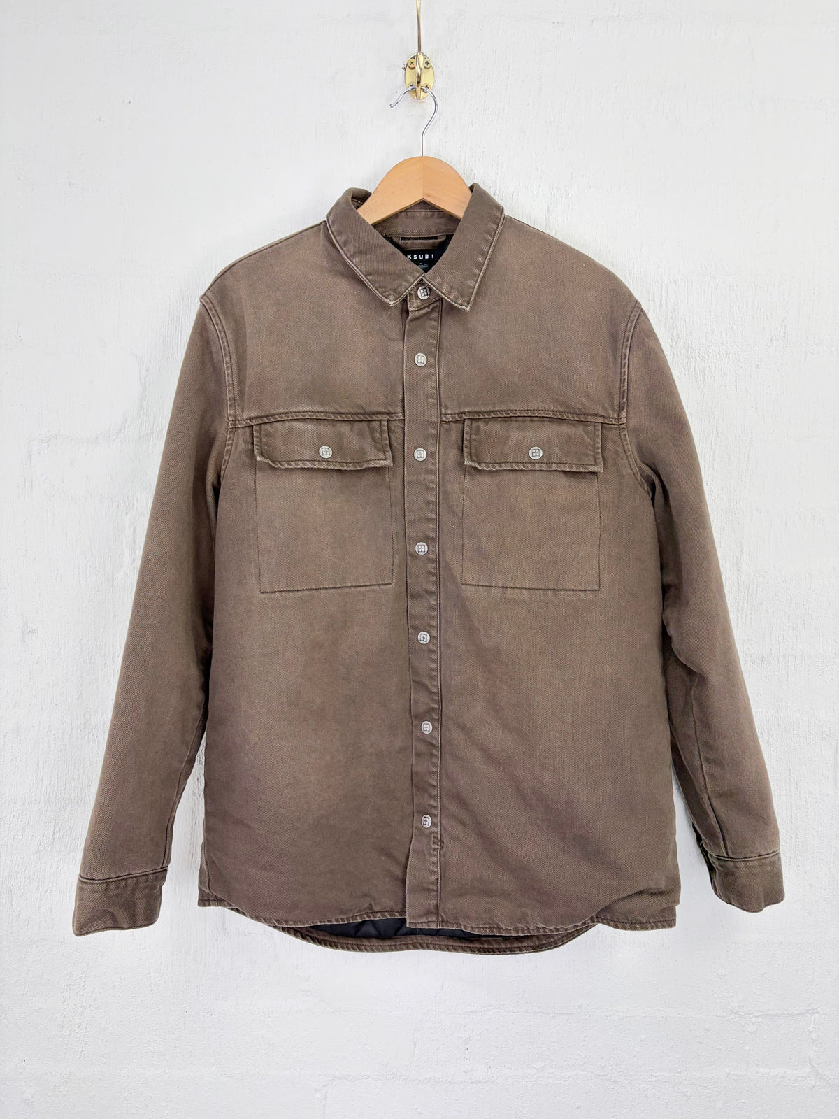 A brown long-sleeve button-up shirt jacket with two chest pockets, hanging on a wooden hanger against a white painted brick wall.