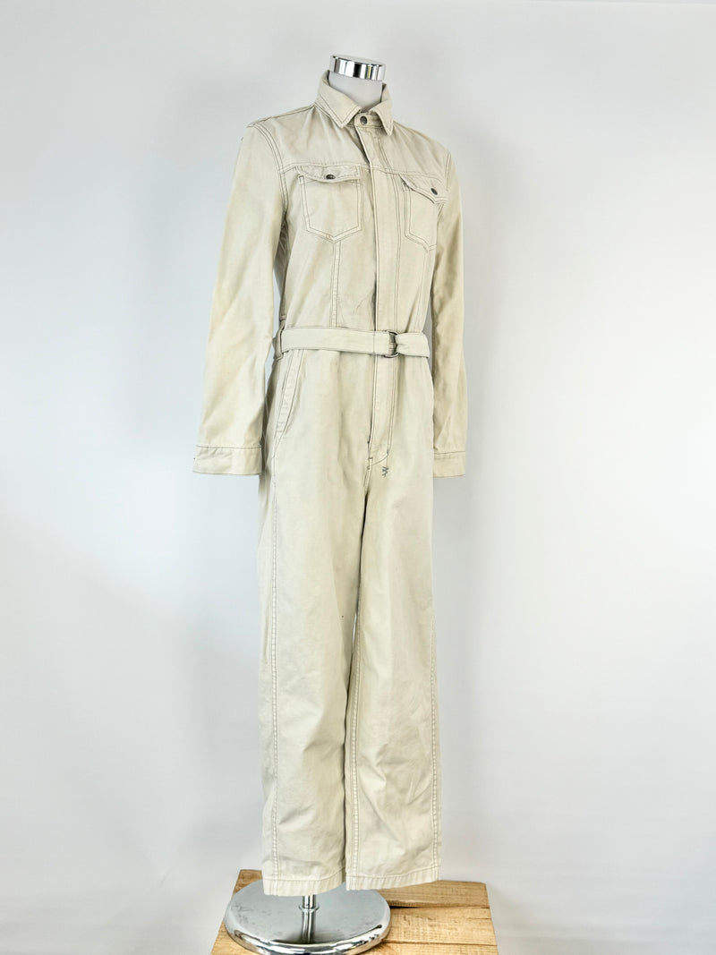A long sleeve beige cotton jumpsuit with multiple pockets and a button-up front, displayed on a mannequin.