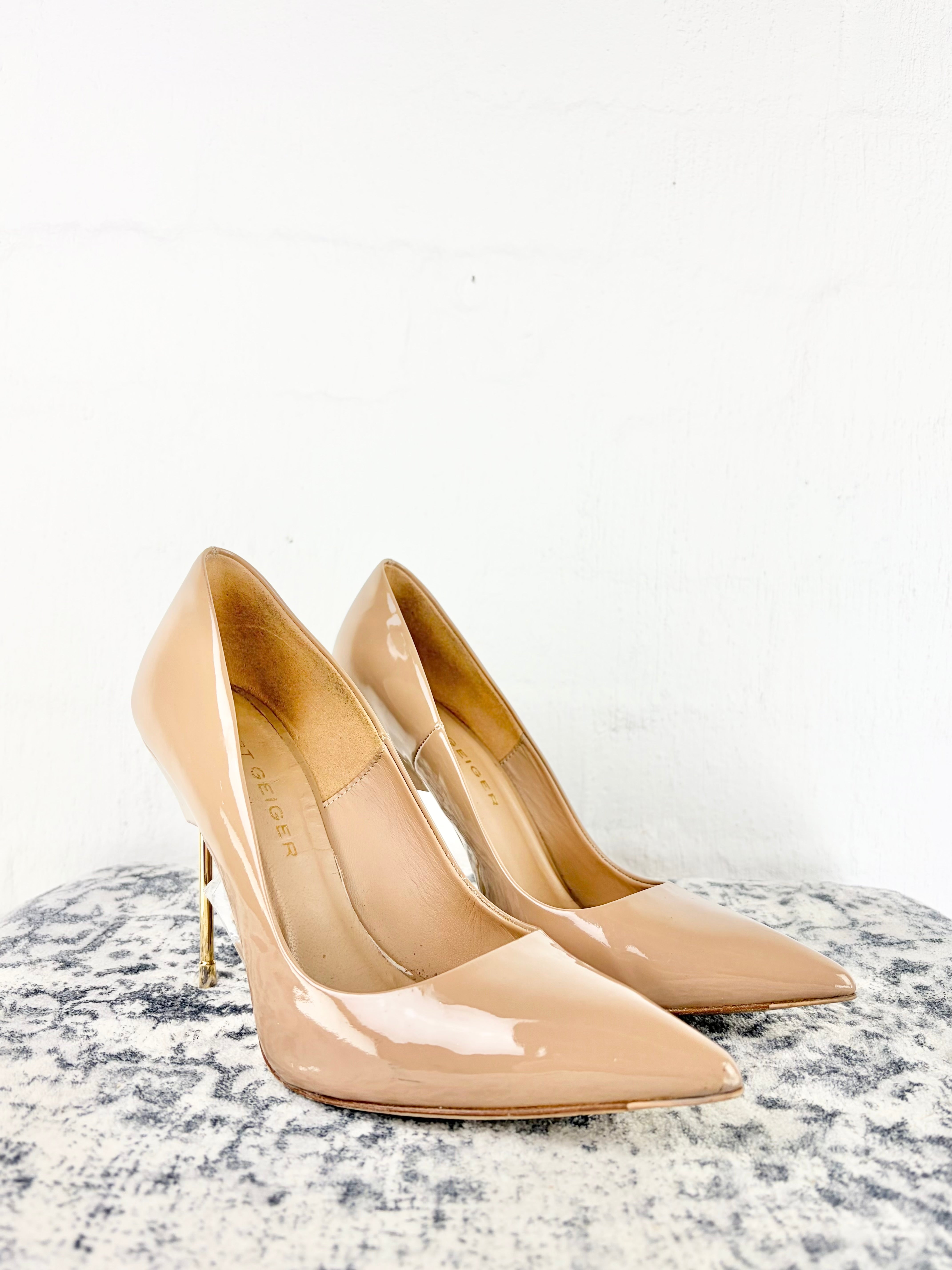 A pair of beige patent leather kitten heel pumps with pointed toes, displayed on a textured gray surface against a white wall.