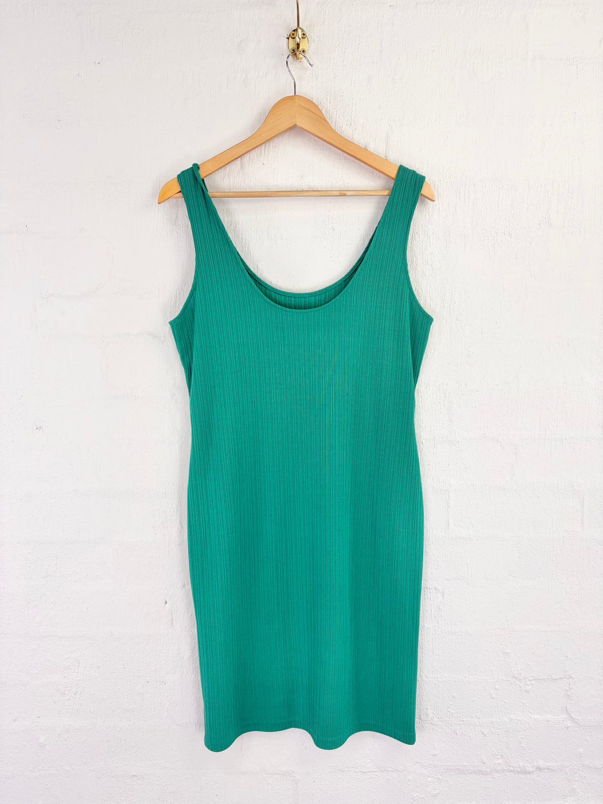 A green ribbed knit sleeveless tank dress hanging on a wooden hanger against a white painted brick wall.