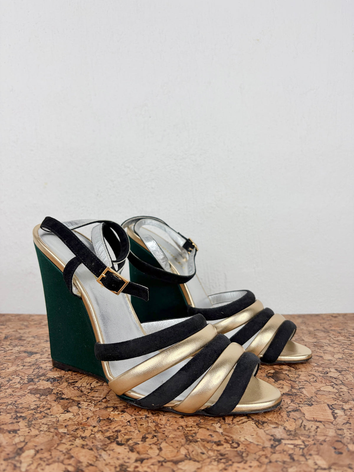 A pair of women's wedge sandals with metallic gold and black straps, featuring a dark green velvet wedge heel and a gold-accented clear toe box, displayed on a cork surface against a white wall.