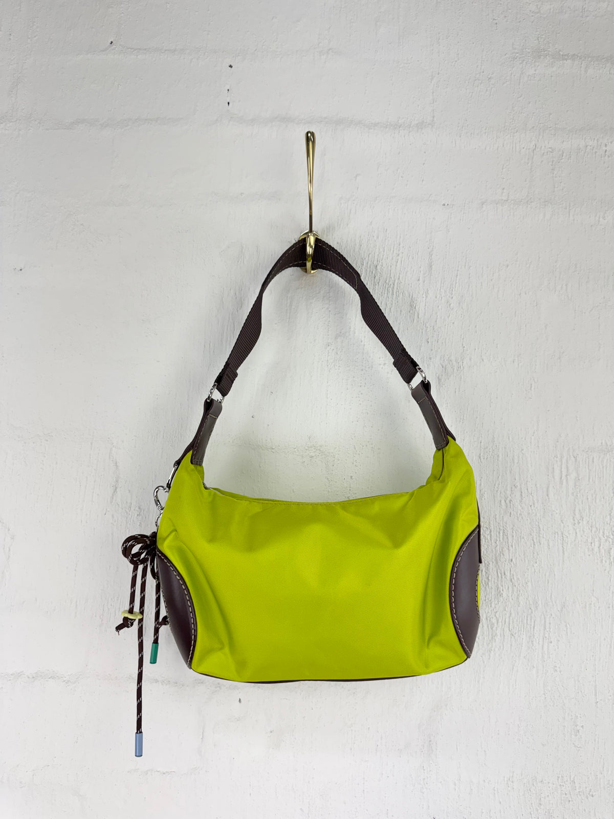 A lime green nylon shoulder bag with dark brown leather trim and a matching adjustable shoulder strap, hanging from a gold hook on a white painted brick wall.