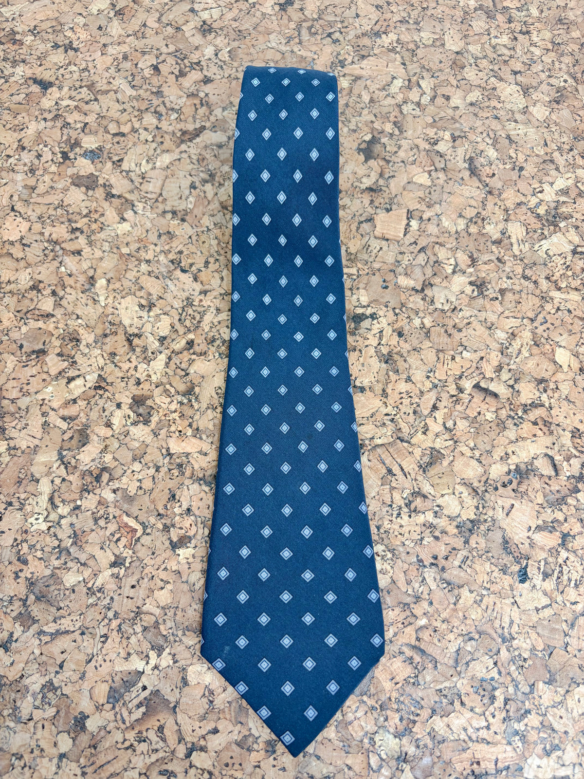 A blue necktie with a small white geometric diamond pattern, laid flat on a textured cork surface.