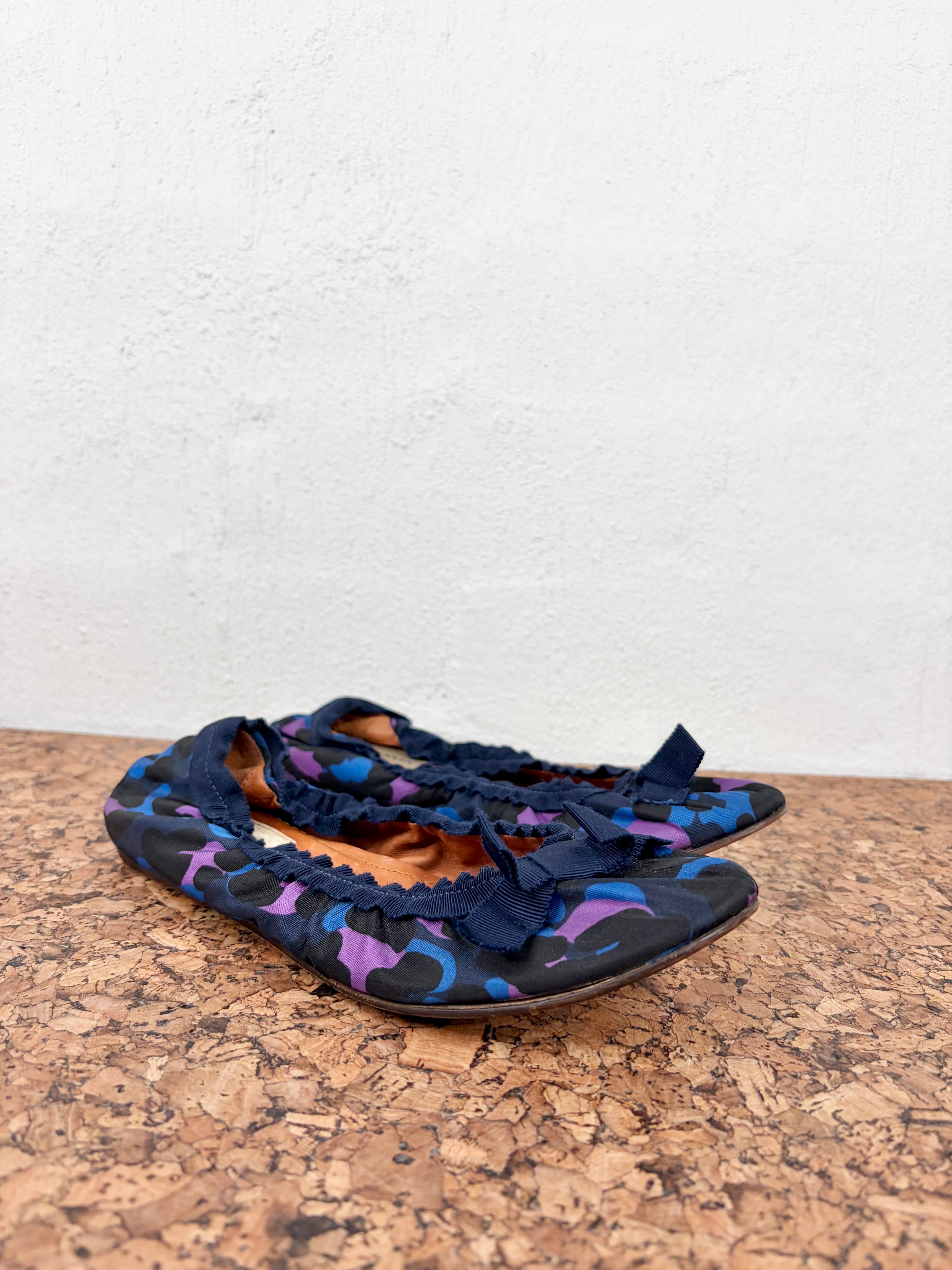 A pair of women's flat shoes with a navy blue base, purple and blue abstract pattern, and dark blue ruffled straps, resting on a cork surface against a white wall.