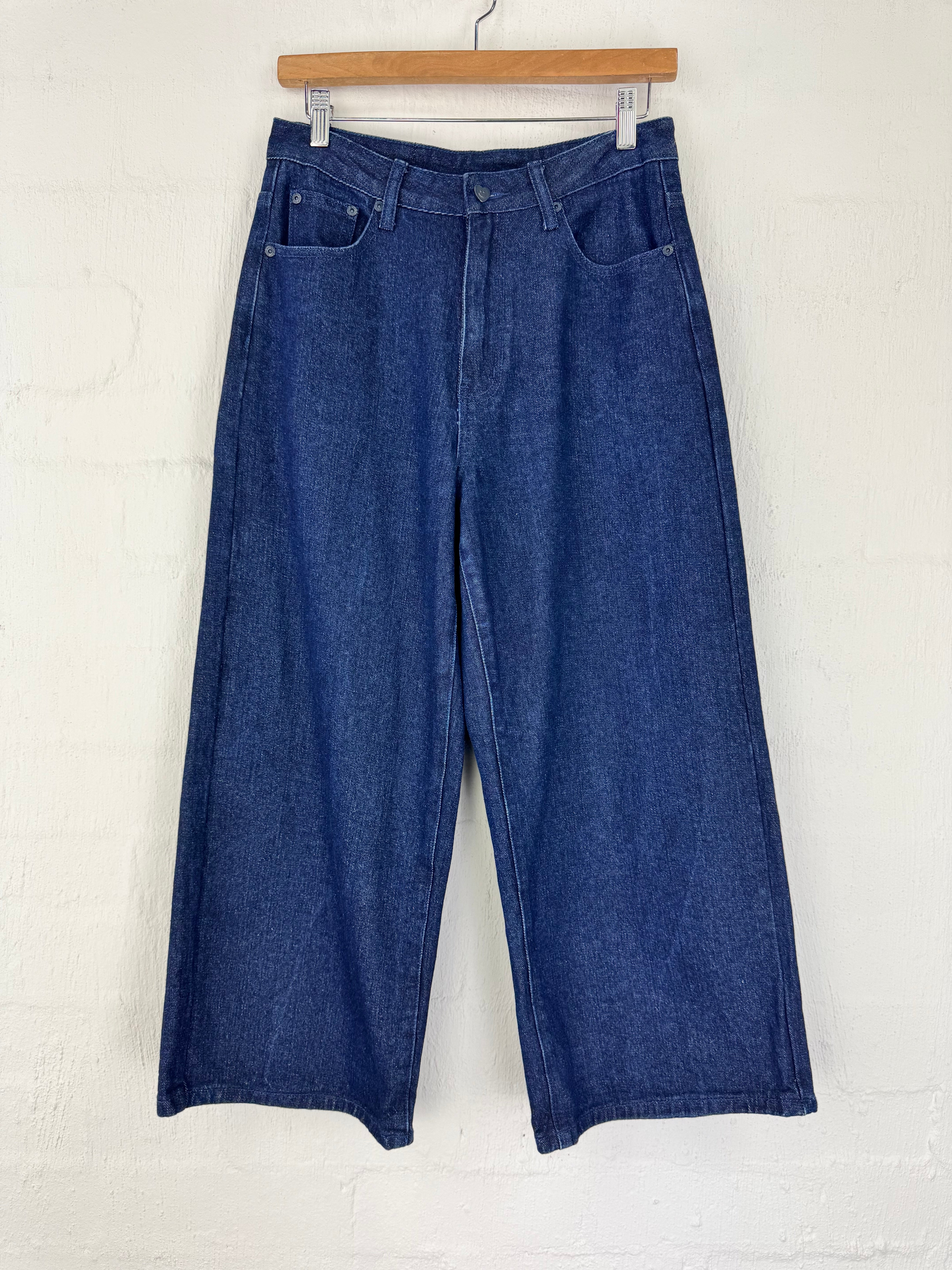 A pair of dark blue wide-leg denim cropped pants hanging from a wooden hanger against a white painted brick wall.