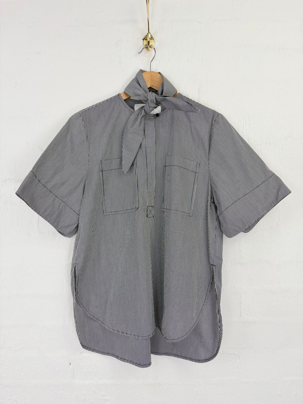 A gray short-sleeve button-up shirt with a tie-neck detail and two chest pockets, hanging on a wooden hanger against a white brick wall.