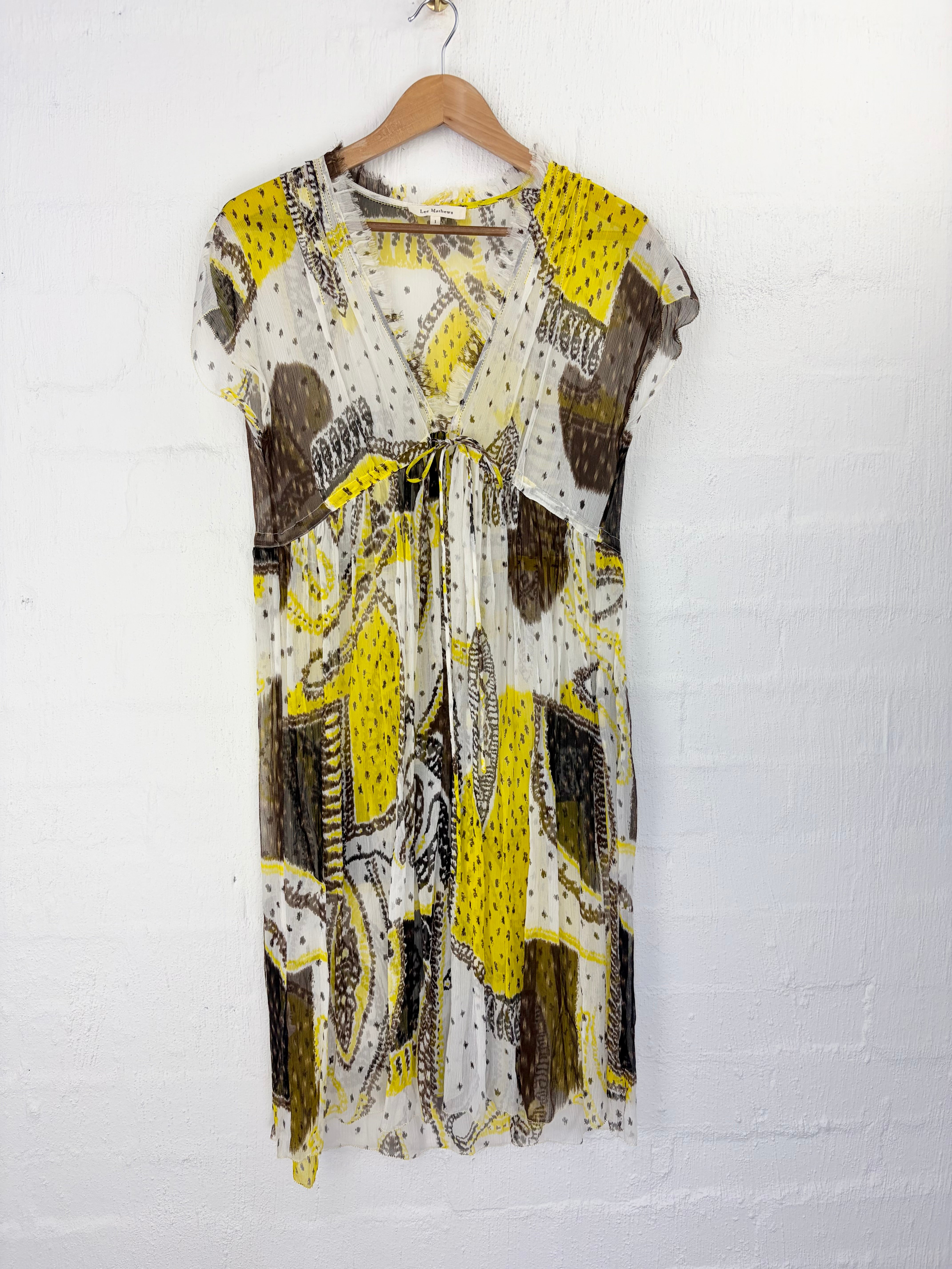 A short-sleeve wrap dress with a tie waist, featuring an abstract patchwork print in yellow, brown, black, and white, hanging on a wooden hanger against a white brick wall.