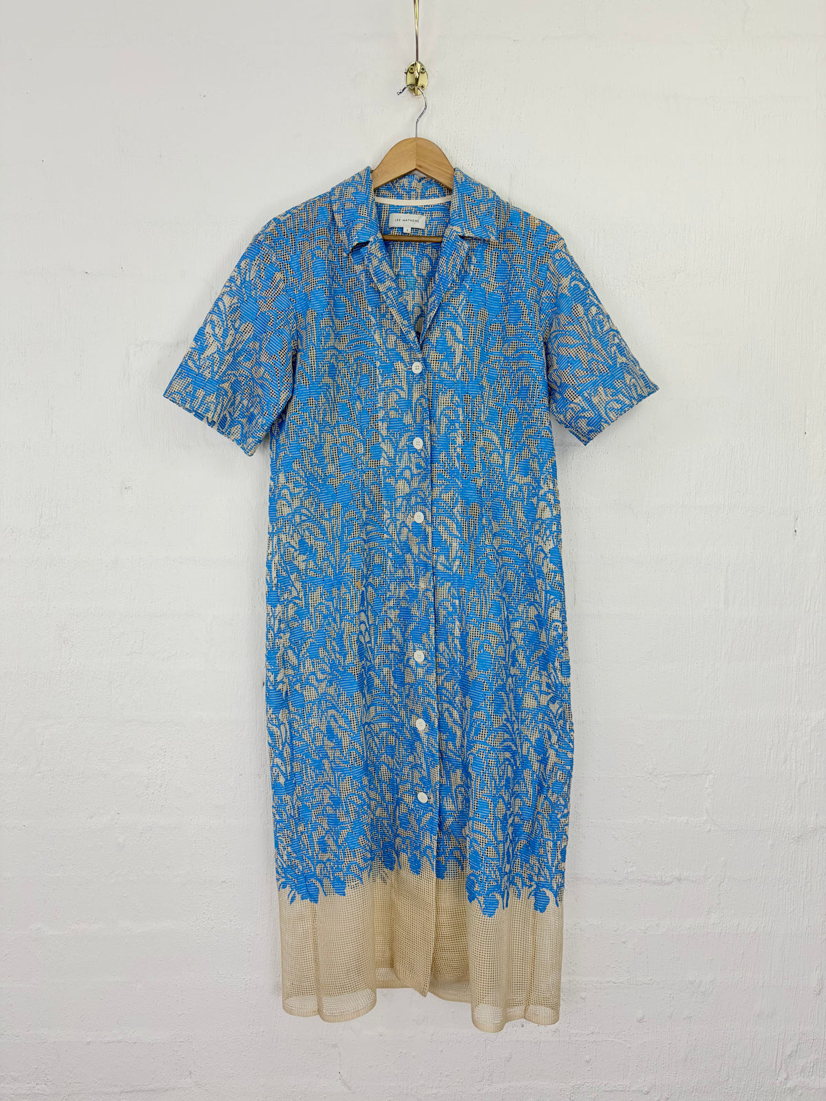A short-sleeve button-front shirt dress with an allover blue and beige floral print, hanging on a wooden hanger against a white painted brick wall.