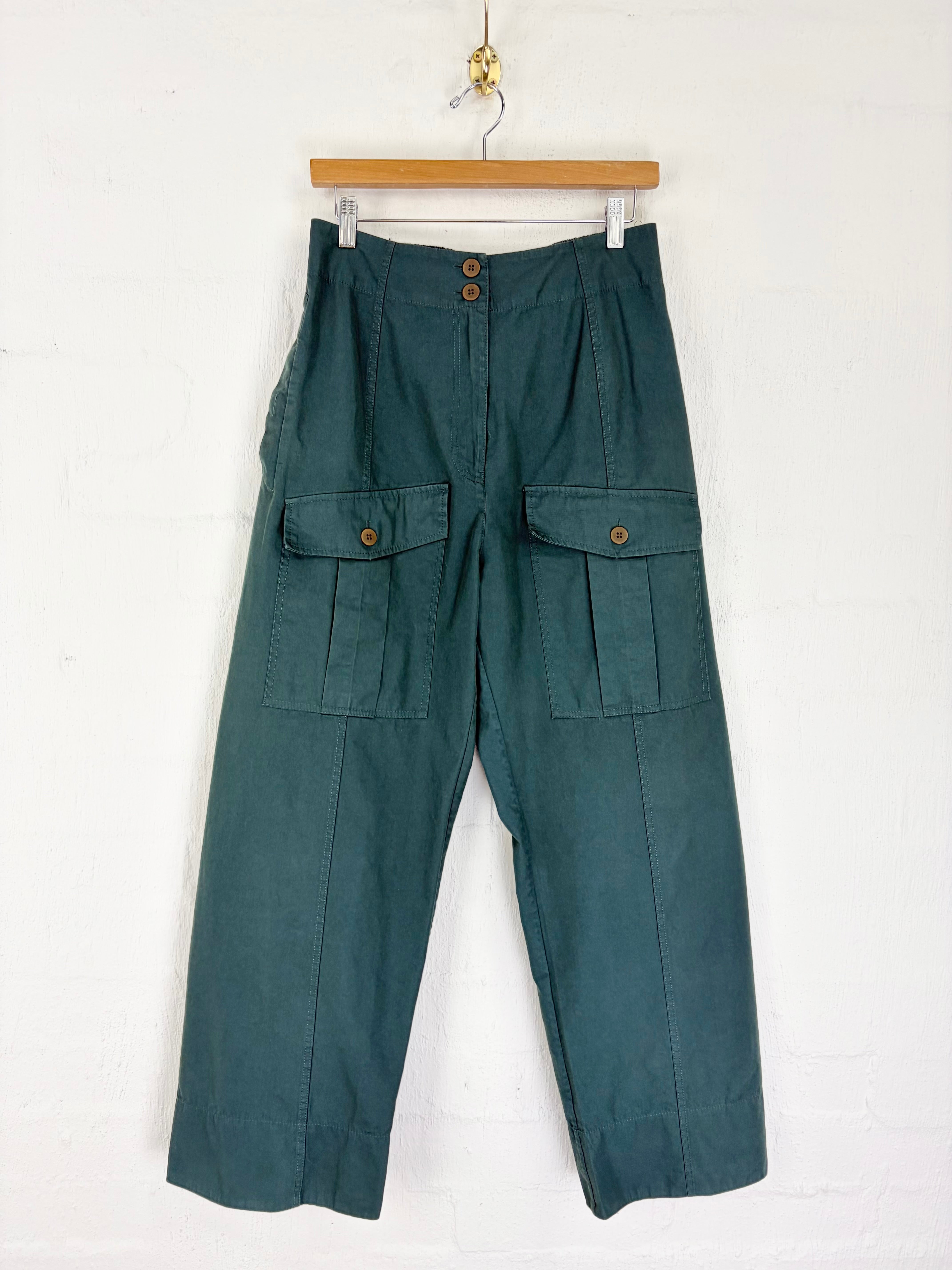 A pair of dark teal green cargo pants hanging from a wooden hanger against a white painted brick wall. The pants feature a high waist, front button closure, and two large flap cargo pockets on the thighs.