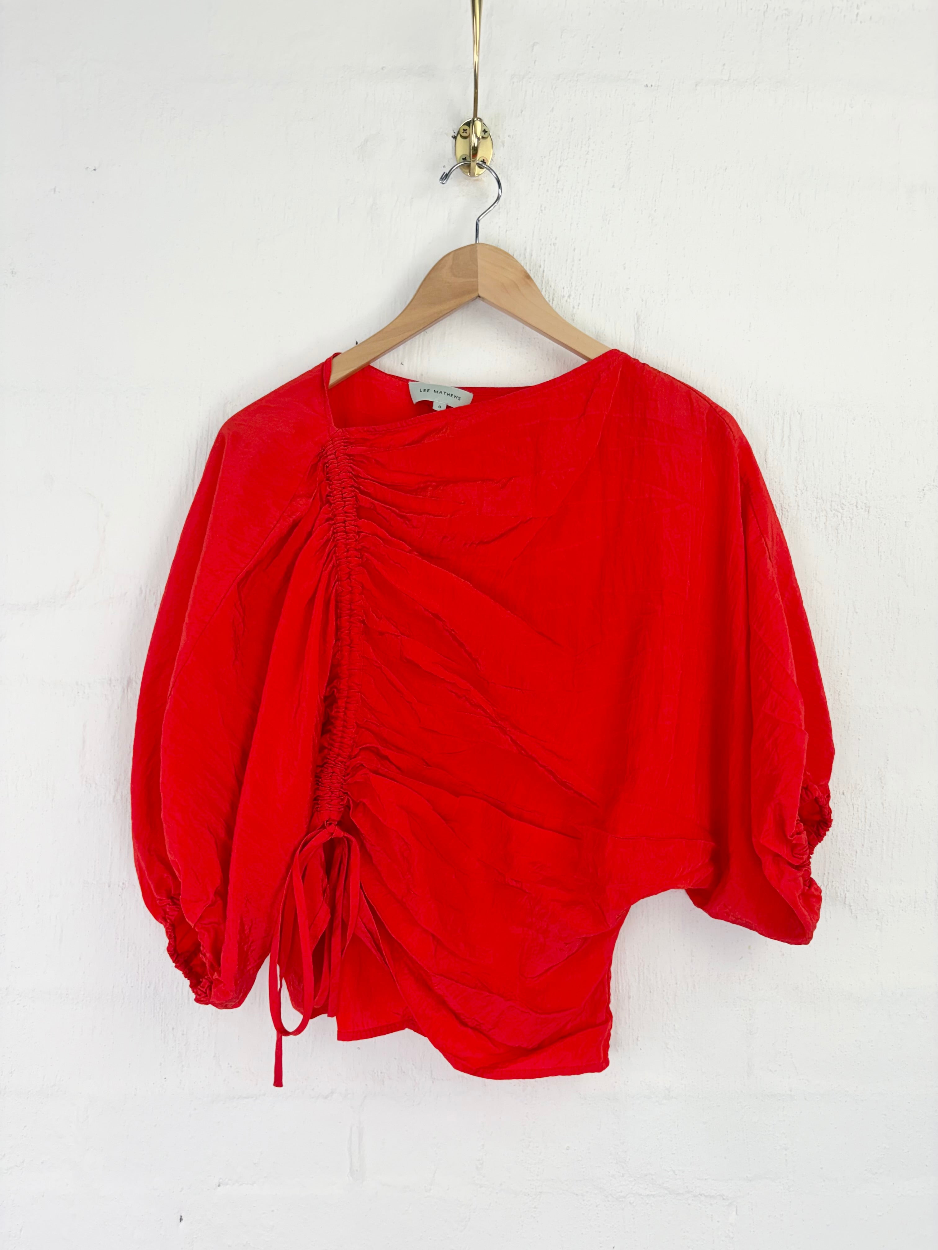 A bright red draped top with balloon sleeves and a side drawstring, hanging on a wooden hanger against a white brick wall.