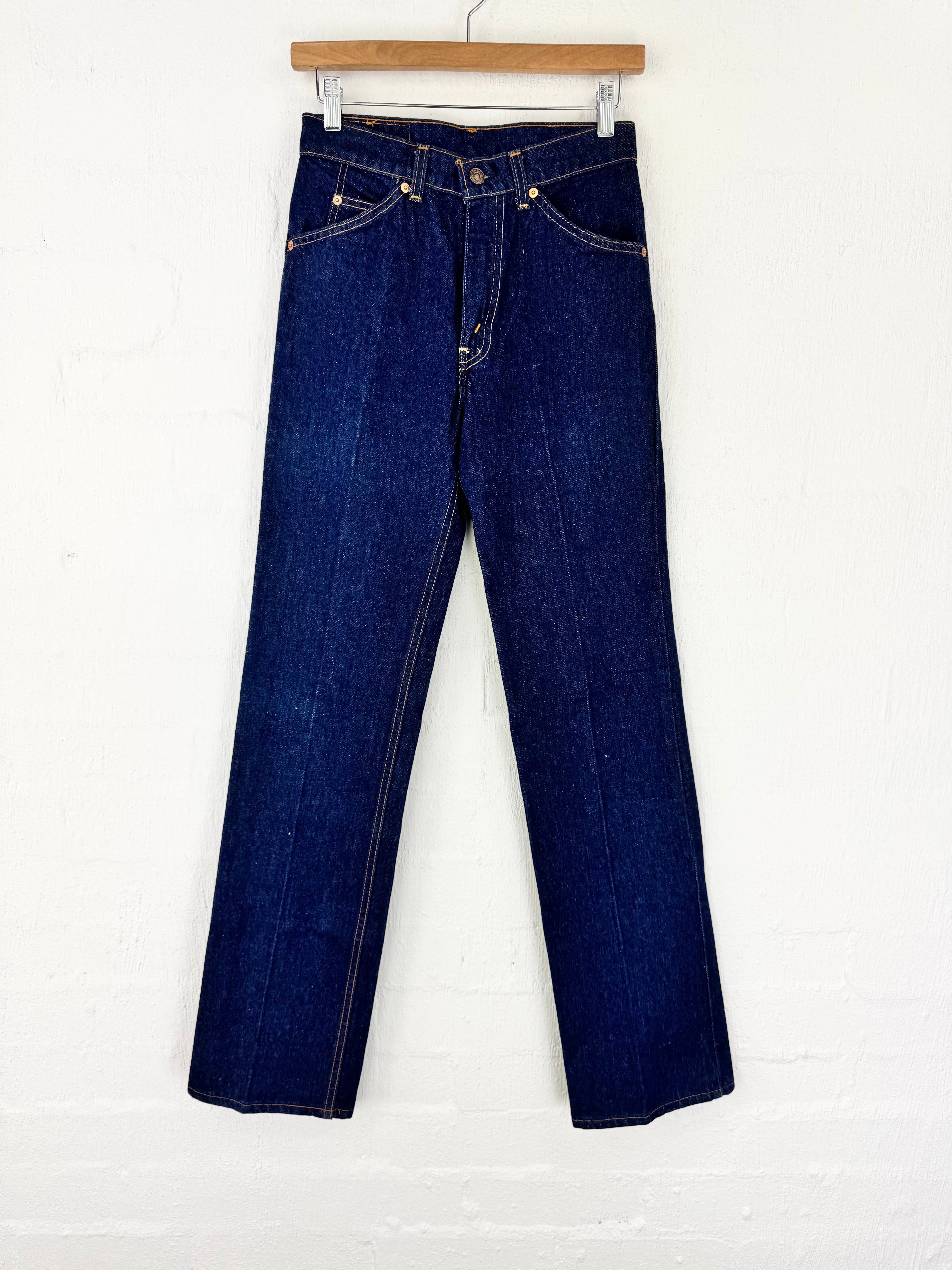 A pair of dark blue denim jeans hanging from a wooden hanger against a white painted brick wall.