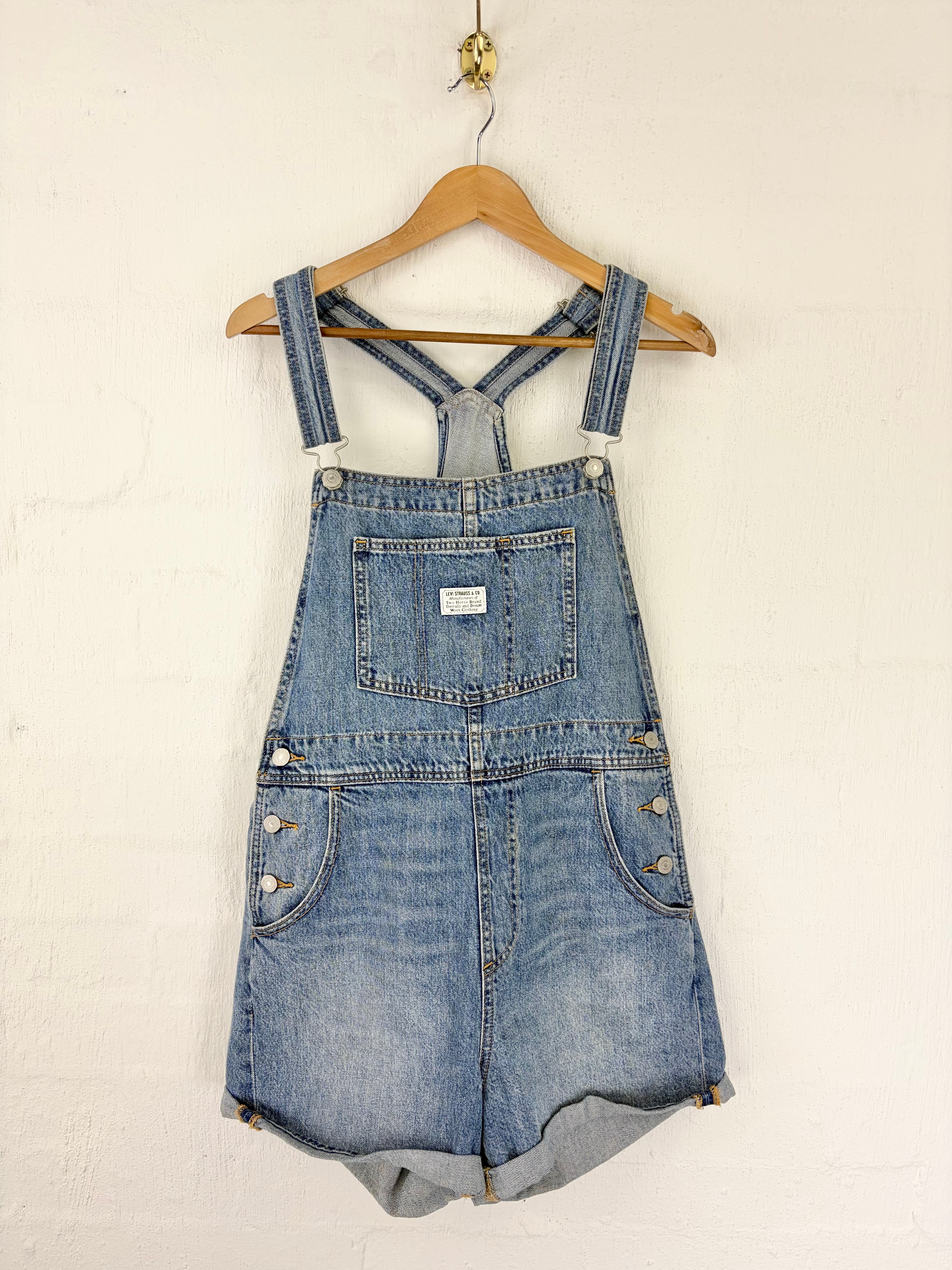Light blue denim overall shorts with adjustable shoulder straps and a cuffed hem, hanging on a wooden hanger against a white painted brick wall.